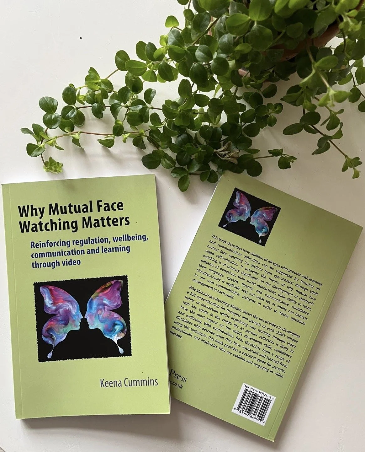 A green book titled "Why Mutual Face Watching Matters" by Keena Cummins, with a related back cover beside it, placed next to a potted green plant with trailing stems on a white surface.