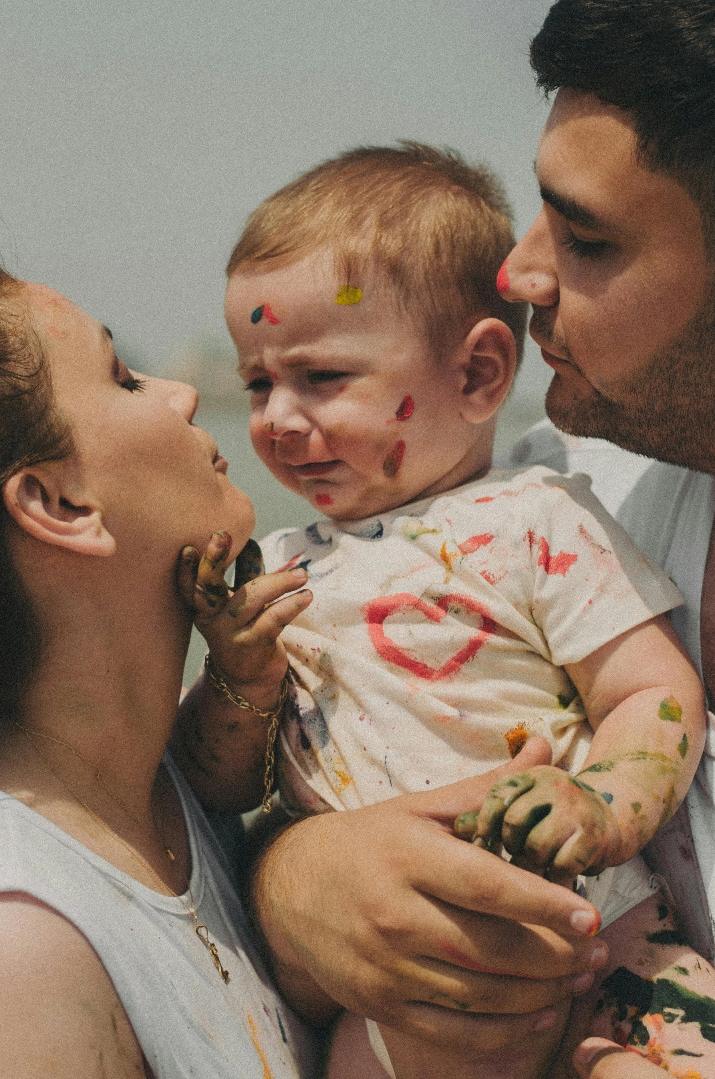 A family holding a crying toddler with paint on their faces and clothes, outdoors on a cloudy day.