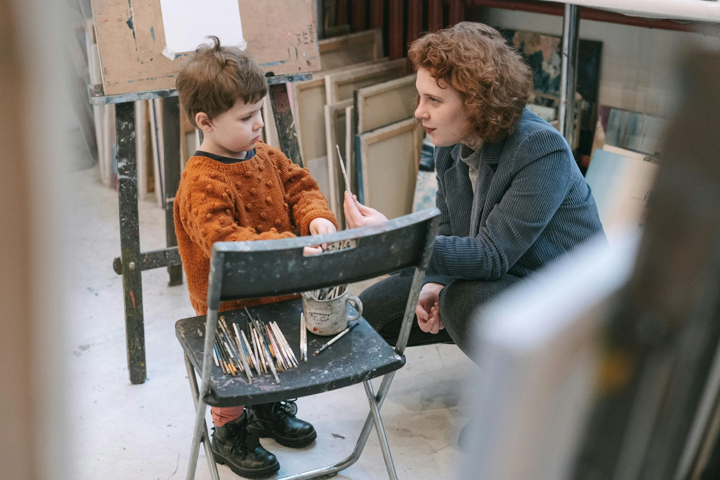 A young boy with brown hair wearing an orange sweater sits at a small table in an art studio with paintbrushes in front of him. A woman with curly hair and a gray blazer is kneeling next to him, holding a paintbrush, and talking to him.