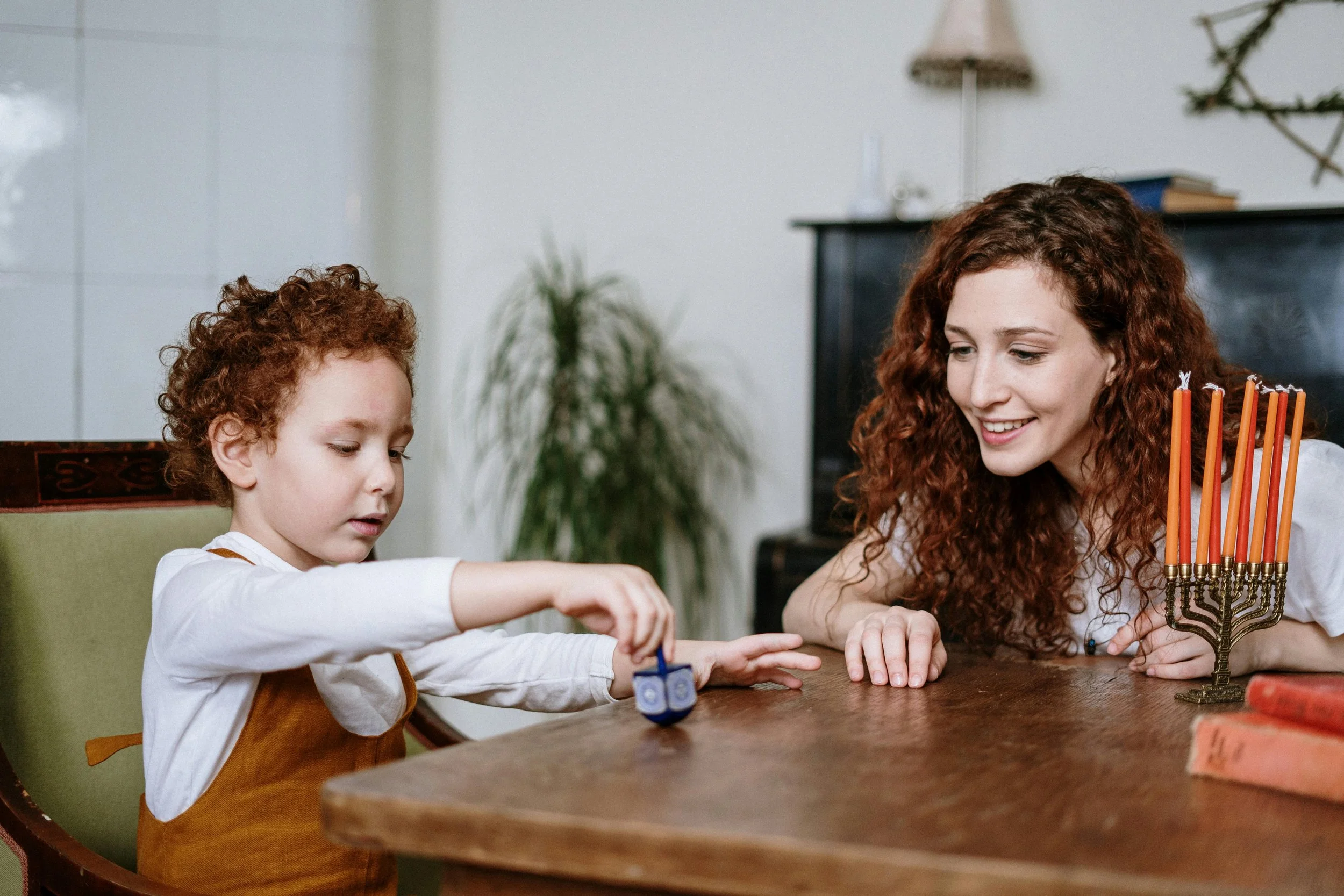 A woman and a young boy are sitting at a wooden table, playing with a small blue toy. The woman has curly red hair and is smiling at the boy, who has similar curly red hair and is focused on the toy. A menorah with candles and some books are on the table.
