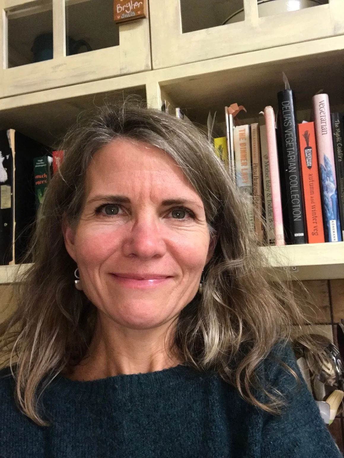 A woman with shoulder-length wavy gray hair smiling in front of a bookshelf filled with cookbooks and a kitchen cabinet.