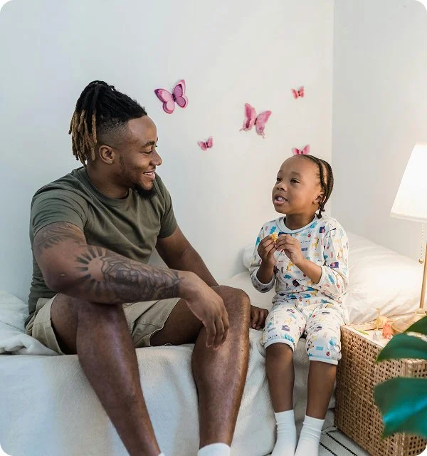A man and a young girl sitting on a bed, smiling and engaging in conversation. The girl is wearing pajama pants and a long-sleeve shirt with colorful patterns, holding a cookie. The man has dreadlocks and tattoos on his arm. Pink butterfly wall decorations are behind them, and a lamp and basket are on the bedside table.