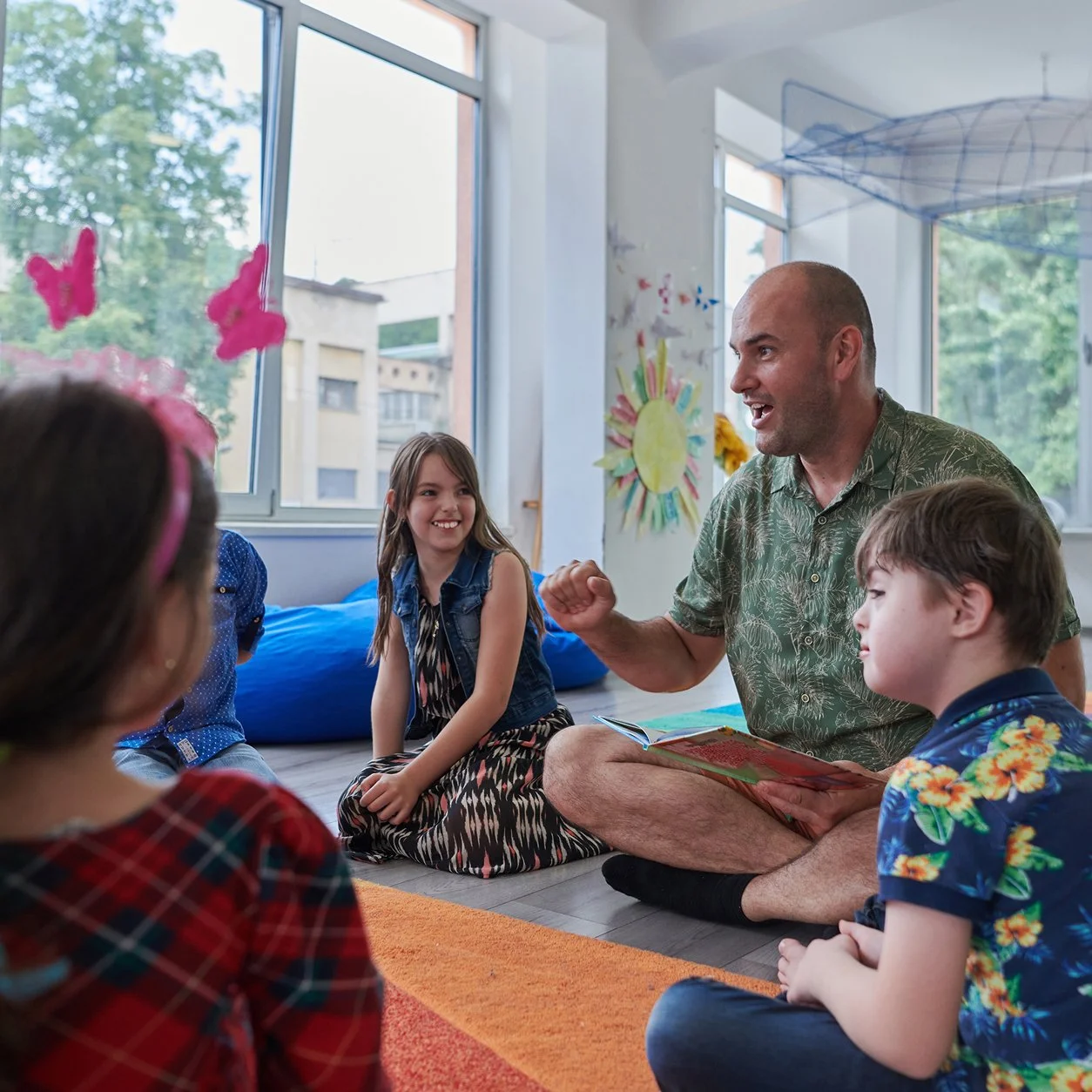 A man reading a picture book to children sitting on the floor in a colorful classroom.