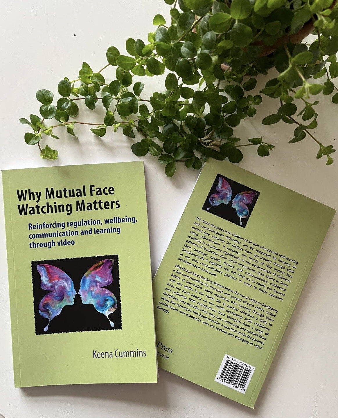A green book titled "Why Mutual Face Watching Matters" by Keena Cummins positioned on a white surface next to a leafy green plant, with the back cover of the book visible.