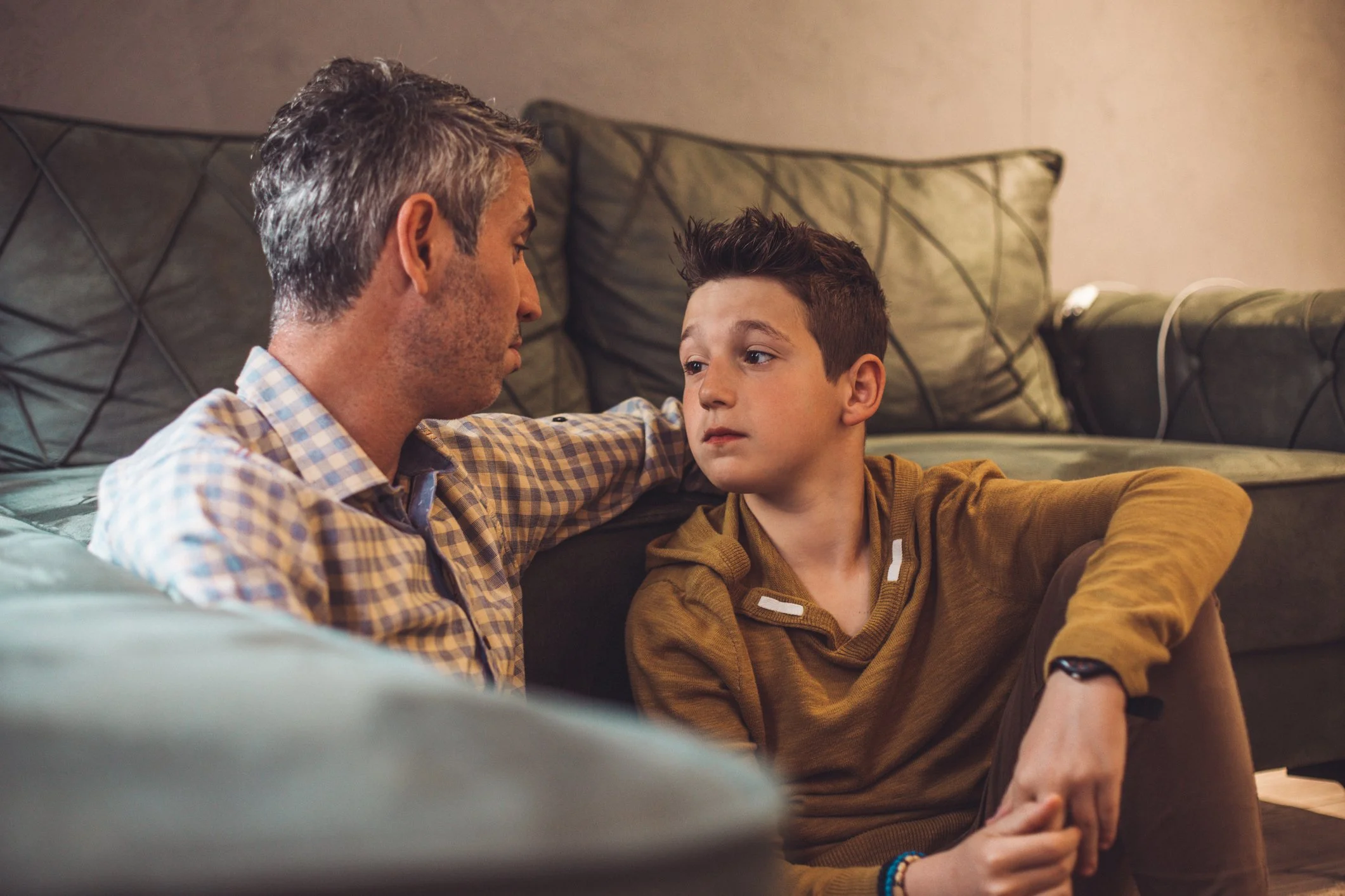 A man with gray hair and a young boy with dark hair sit on the floor of a living room, facing each other with a serious or contemplative expression.