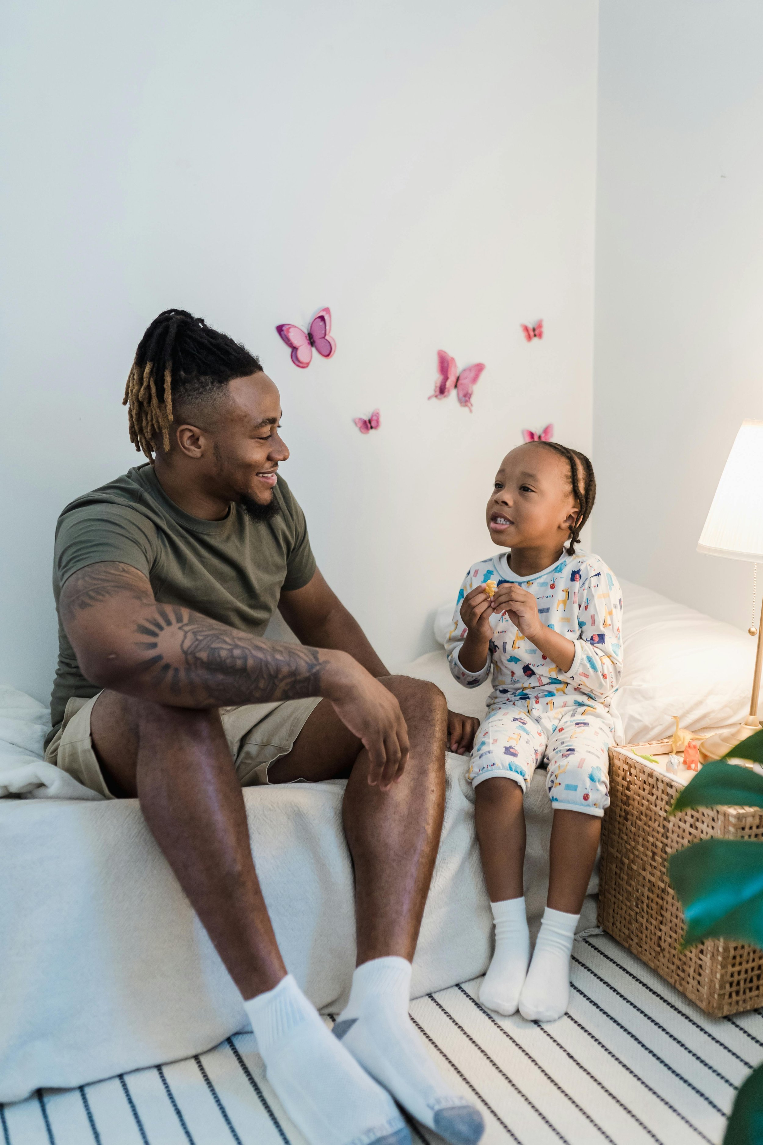 A man and a young girl sitting on a bed, smiling and talking. The girl is holding a snack, wearing pajamas with colorful patterns, and the man is wearing a green t-shirt and beige shorts. Pink butterfly decorations are on the wall behind them.