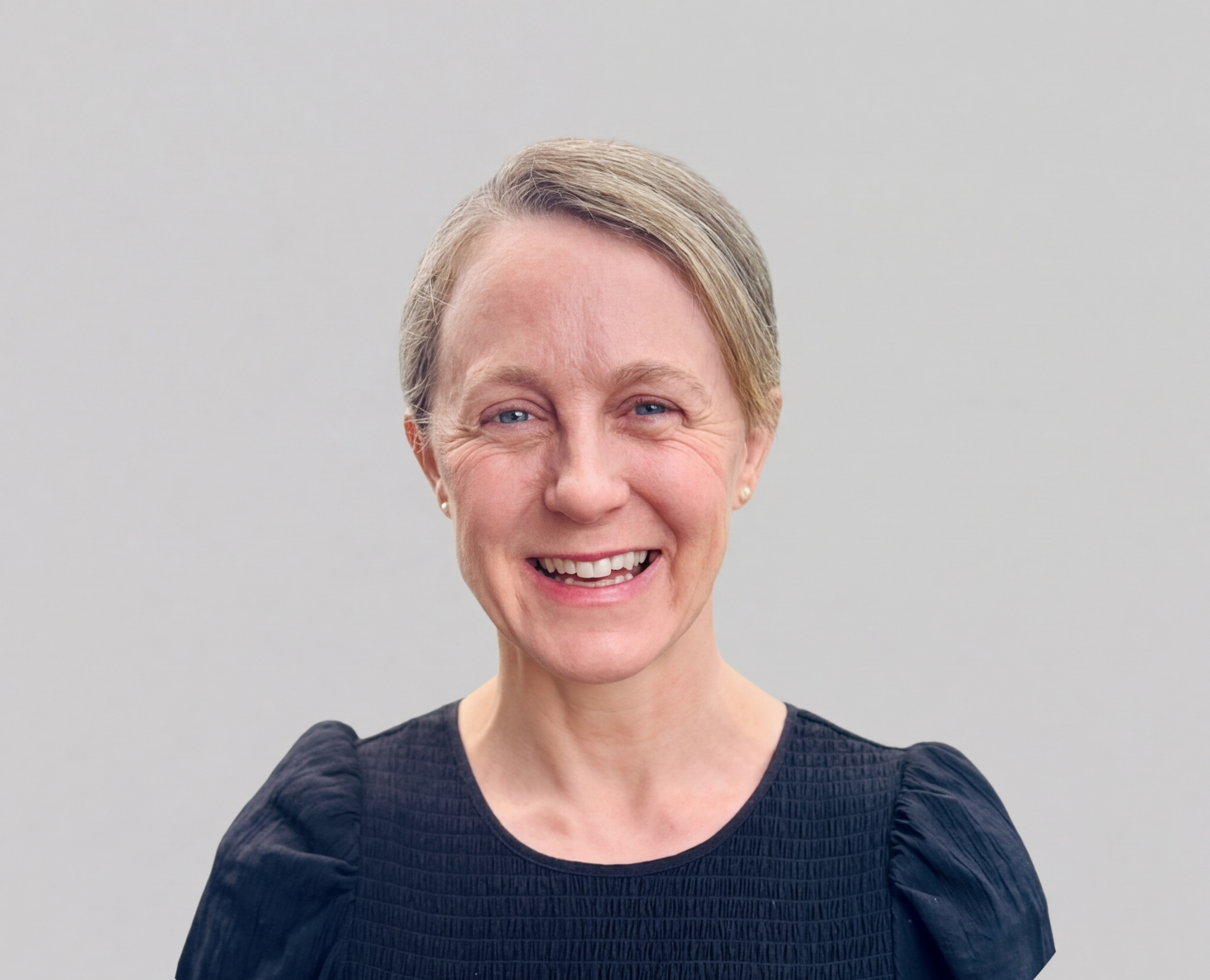Smiling middle-aged woman with short blonde hair, wearing a black top, standing against a plain gray background.
