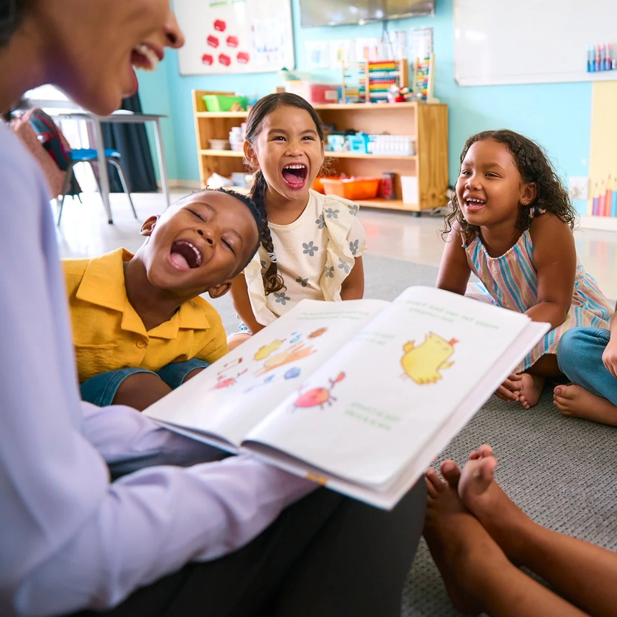 Children sitting on the floor and laughing while an adult reads a colorful picture book to them in a classroom.