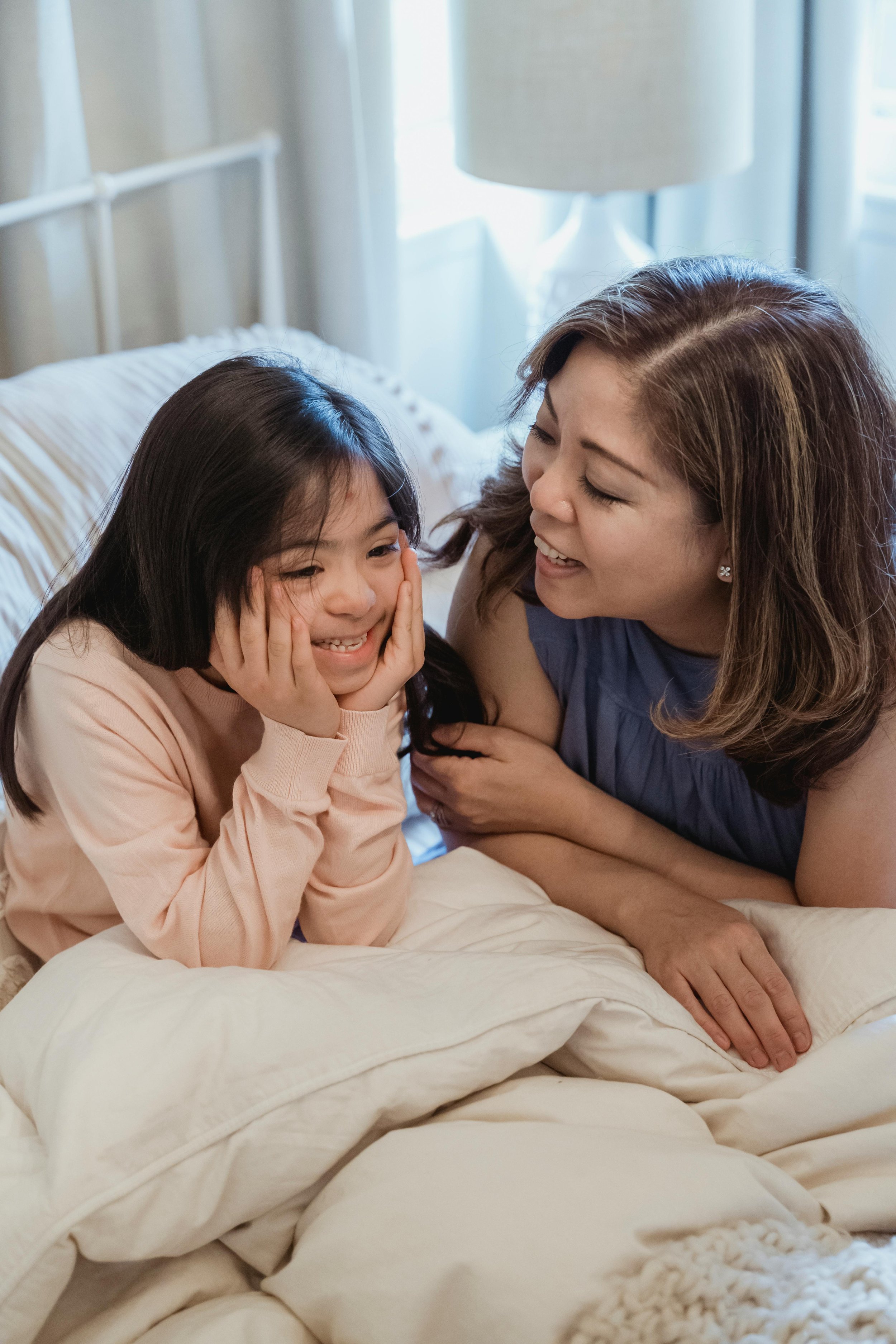 A woman and a young girl are lying on a bed together, smiling and playing. The woman is looking at the girl with a joyful expression while the girl is resting her chin on her hands with a happy face, in a cozy bedroom with natural light.