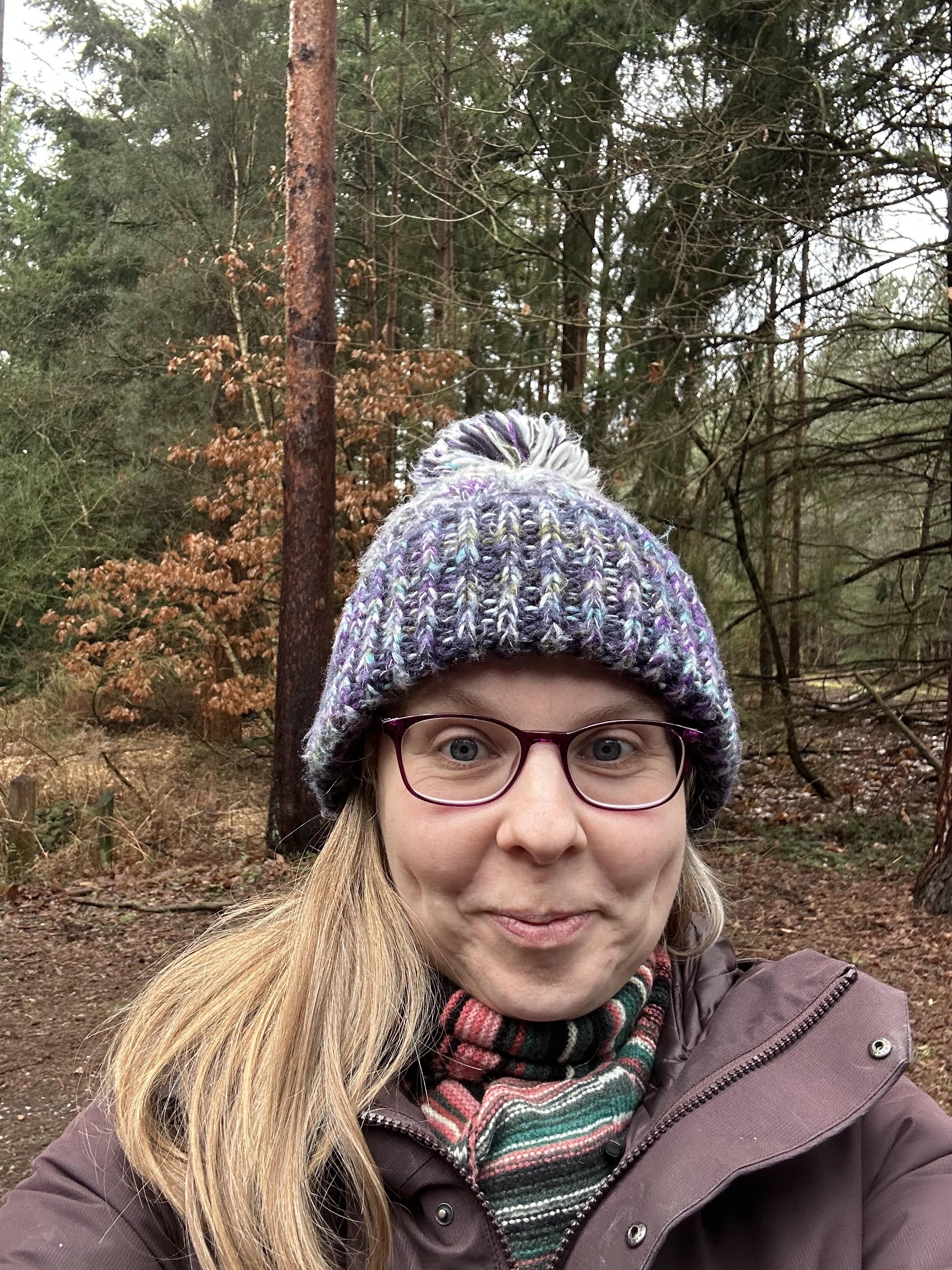 A woman with glasses, wearing a colorful striped scarf and a knitted winter hat with pom-pom, taking a selfie in a forest with tall trees and brown leaves in the background.