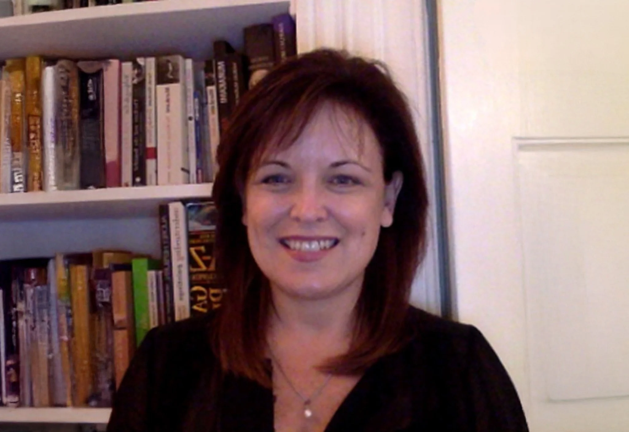 A woman with shoulder-length brown hair, smiling, standing in front of a bookshelf filled with books.