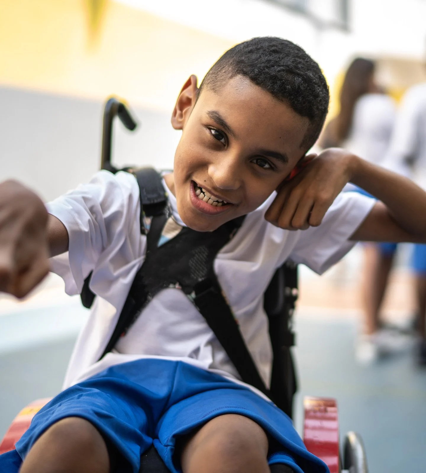 Smiling young boy in a wheelchair with a black harness, wearing a white shirt and blue shorts, in an indoor setting with other children in the background.