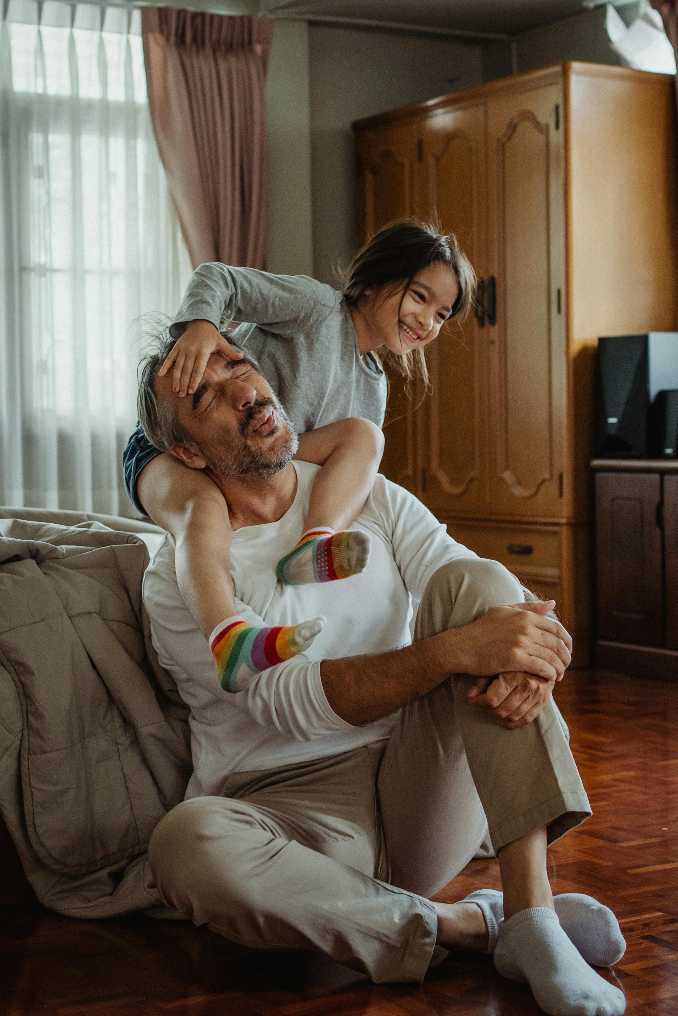 A young girl and an older man, likely her grandfather, playing on the floor of a living room. The girl is sitting on the man's shoulders, holding his head, and both are smiling and laughing. The room has a large window with curtains, a wooden cabinet, and a television.