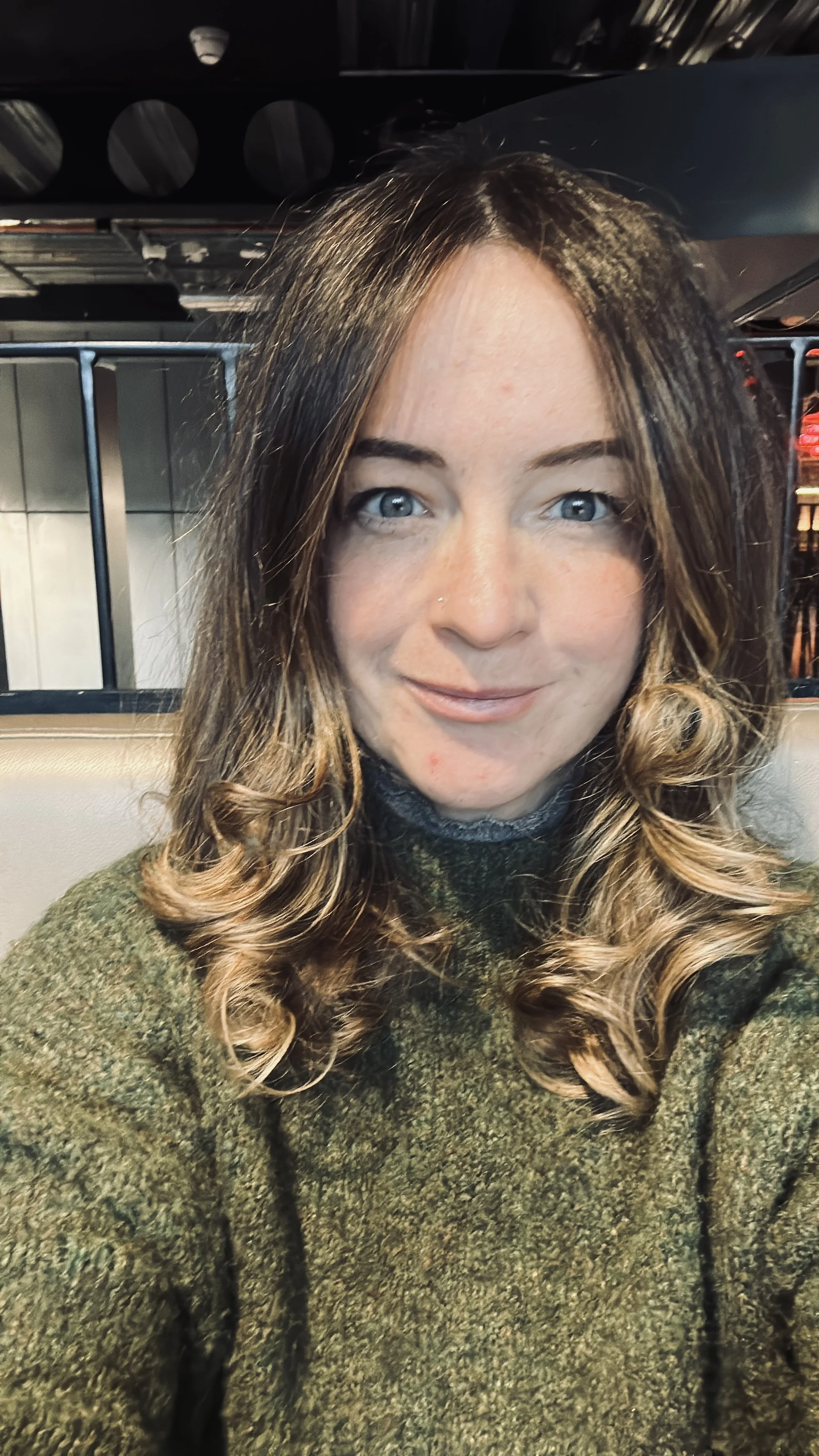 Close-up of a woman with long, curly hair, blue eyes, wearing a green sweater, sitting indoors.