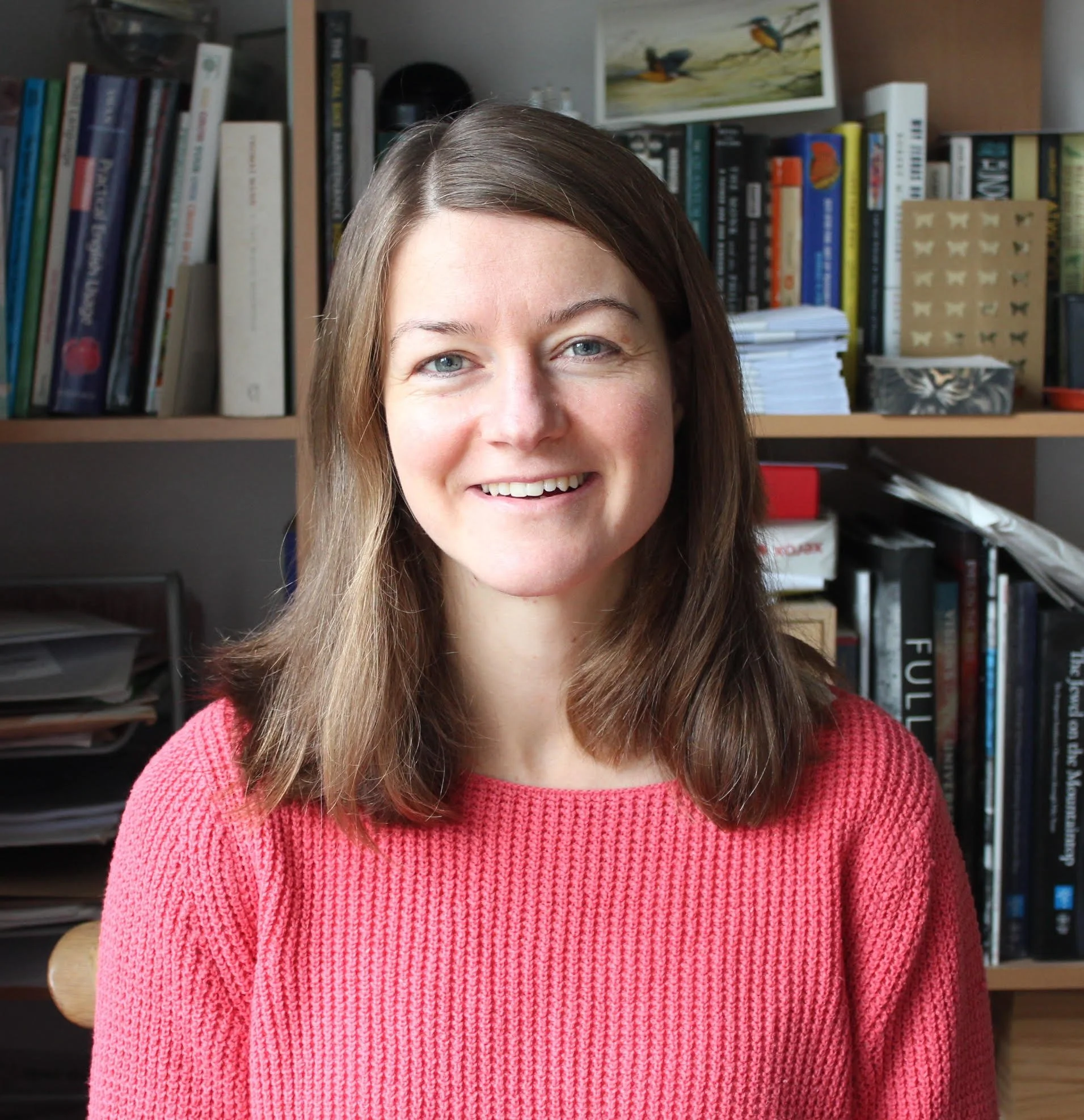 A woman with shoulder-length brown hair and blue eyes, smiling, wearing a pink sweater, sitting in front of a bookshelf filled with books and papers.
