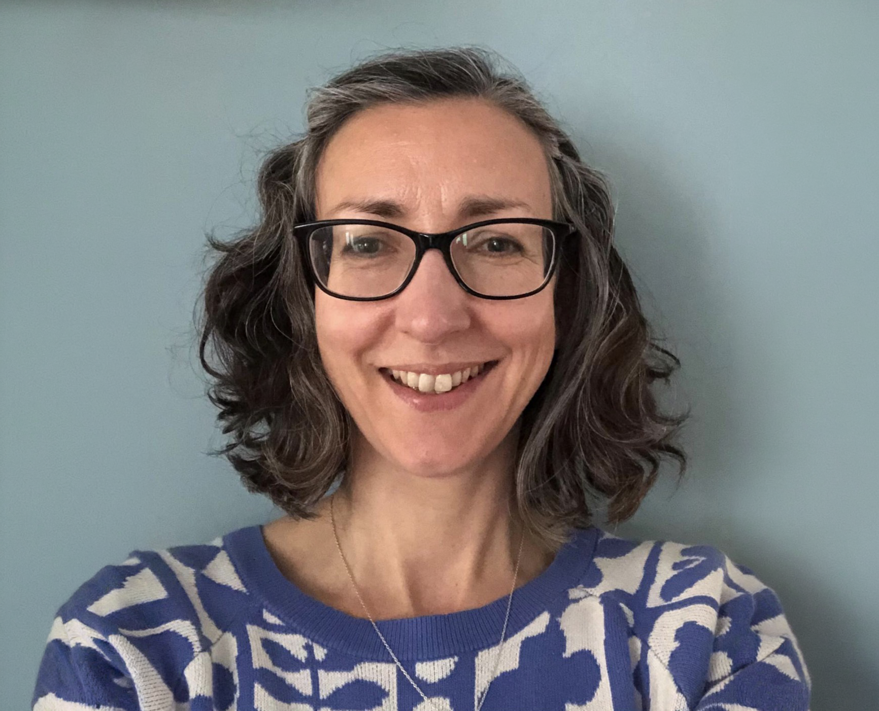 A woman with curly gray hair, glasses, and a blue and white patterned top smiling at a camera, standing against a light blue wall.