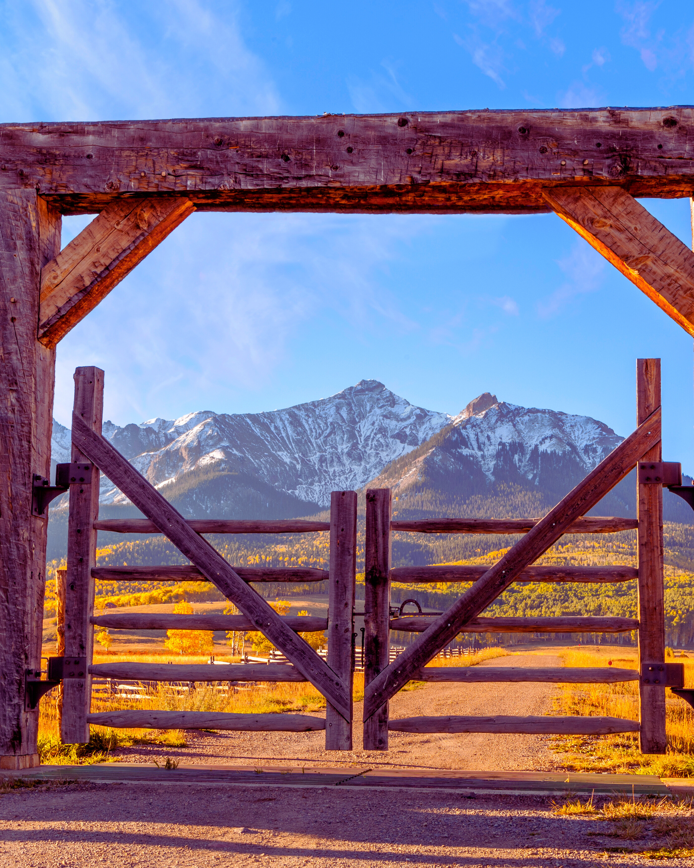 Wooden gate opening to a scenic mountain view with snow-capped peaks, green forest, and autumn trees in the distance, under a blue sky.