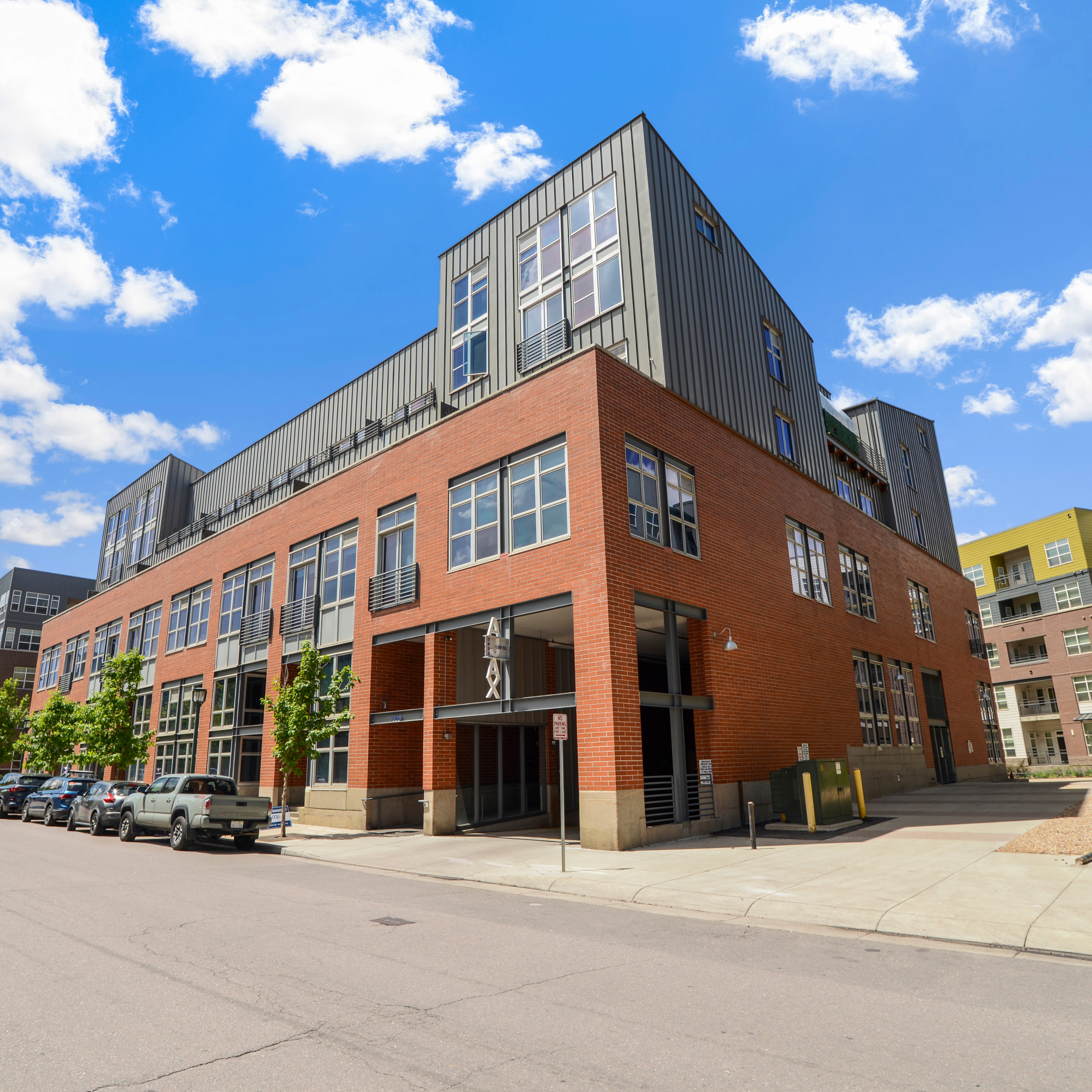 Modern multi-story residential or commercial building with brick and metal exterior, large windows, and parking area in front under a blue sky with clouds.