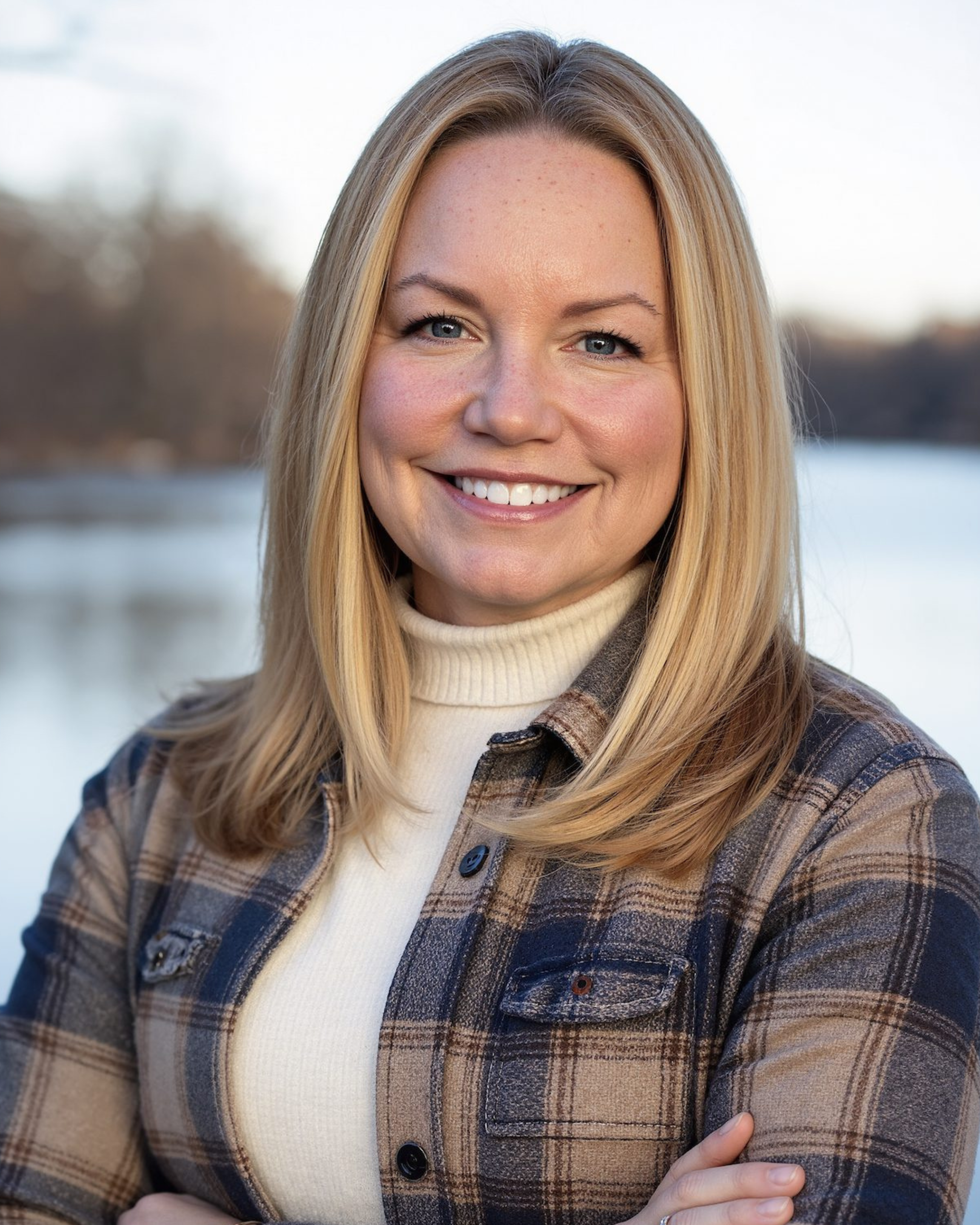 A woman with blonde hair smiling outdoors near a body of water during late afternoon or early evening, wearing a plaid jacket and cream turtleneck.