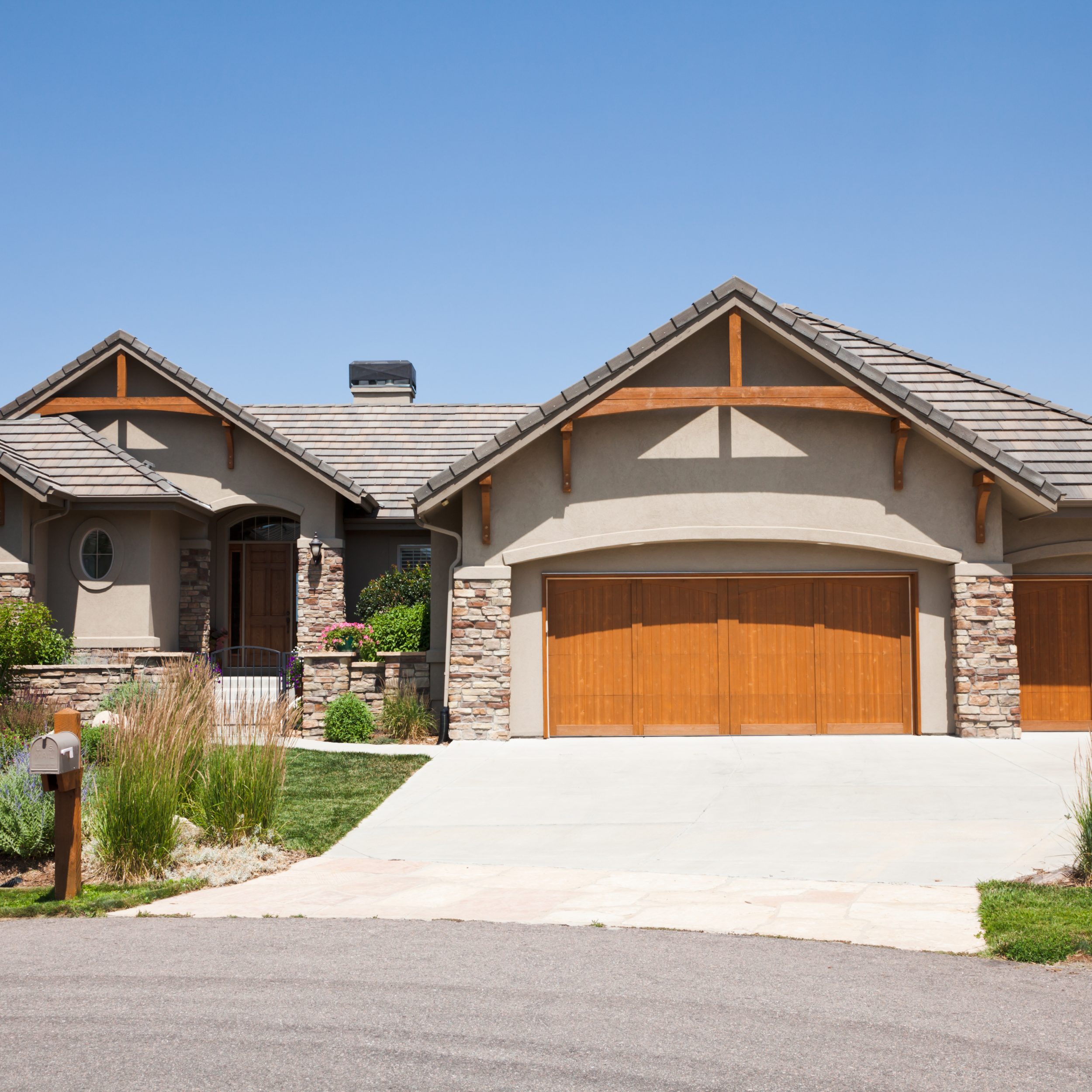 Front view of a modern house with a stone and stucco exterior, wooden garage doors, and landscaped yard on a sunny day.