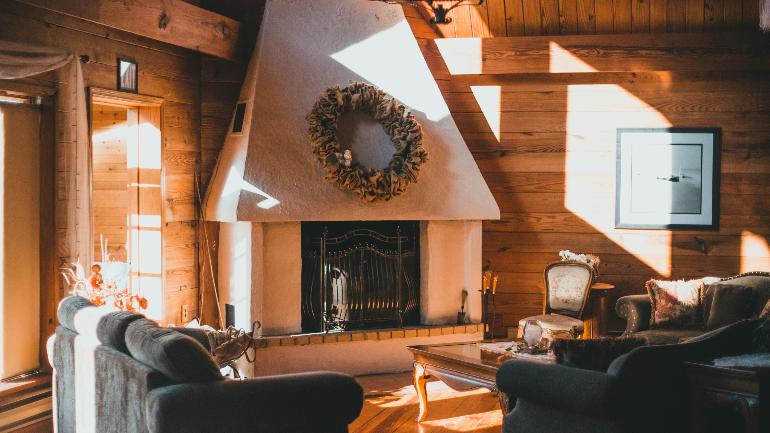 A cozy living room with wooden walls and ceiling, featuring a fireplace with a wreath hanging above it, a framed picture on the wall, a sofa, and a chair. Sunlight streams through the window, casting shadows inside.
