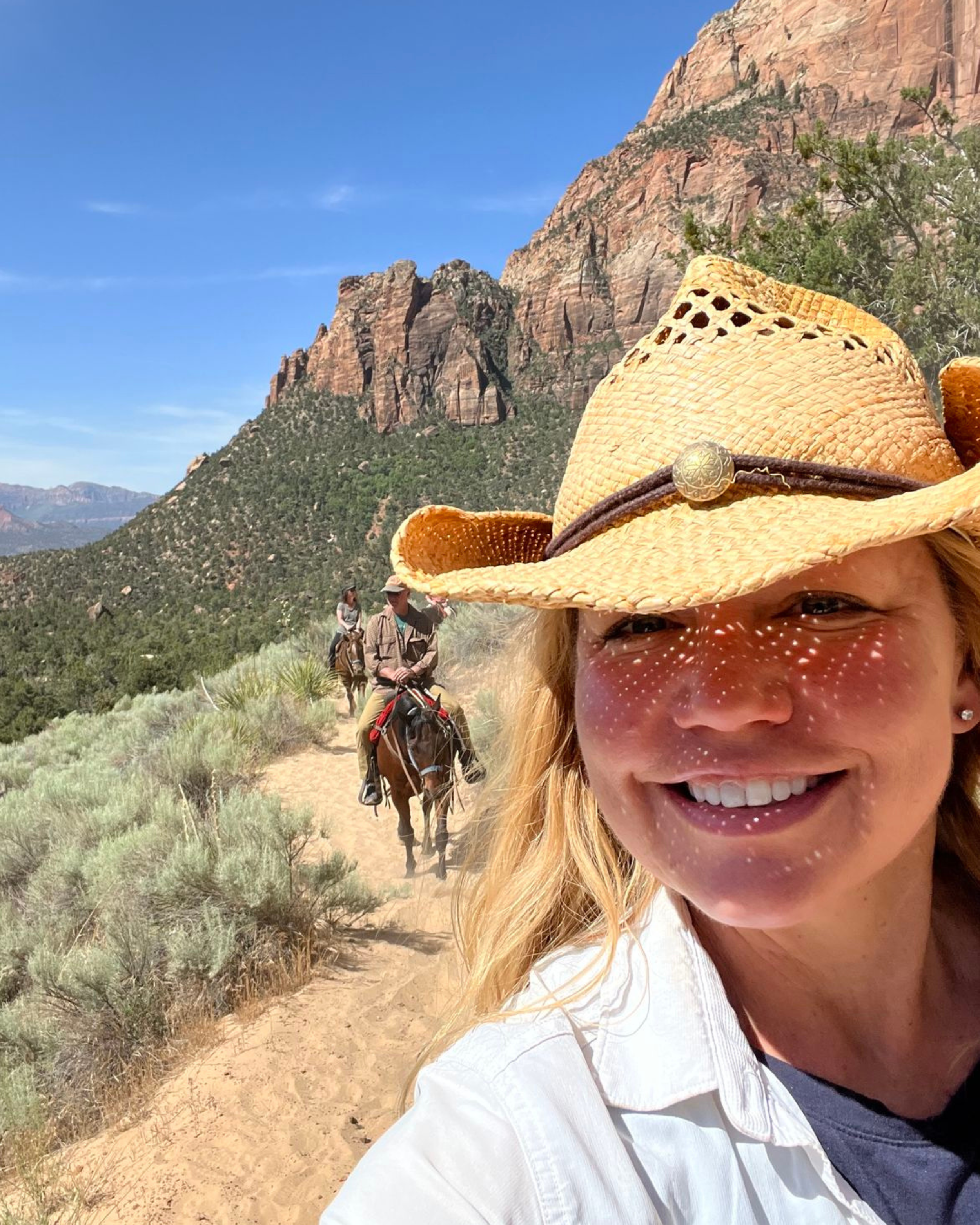 A woman smiling with shadow of a straw hat on her face taking a selfie, with two people riding horses on a sandy trail in a mountainous desert landscape with red rock formations and blue sky in the background.