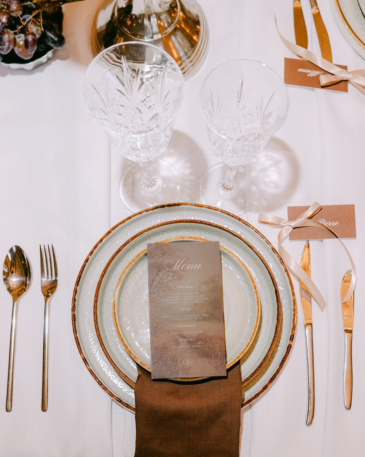 Table élégante avec assiettes en porcelaine or, couteaux, fourchette, cuillères en métal doré, verres en cristal taillé, menu et marque-place avec ruban beige, décor de fête ou mariage.