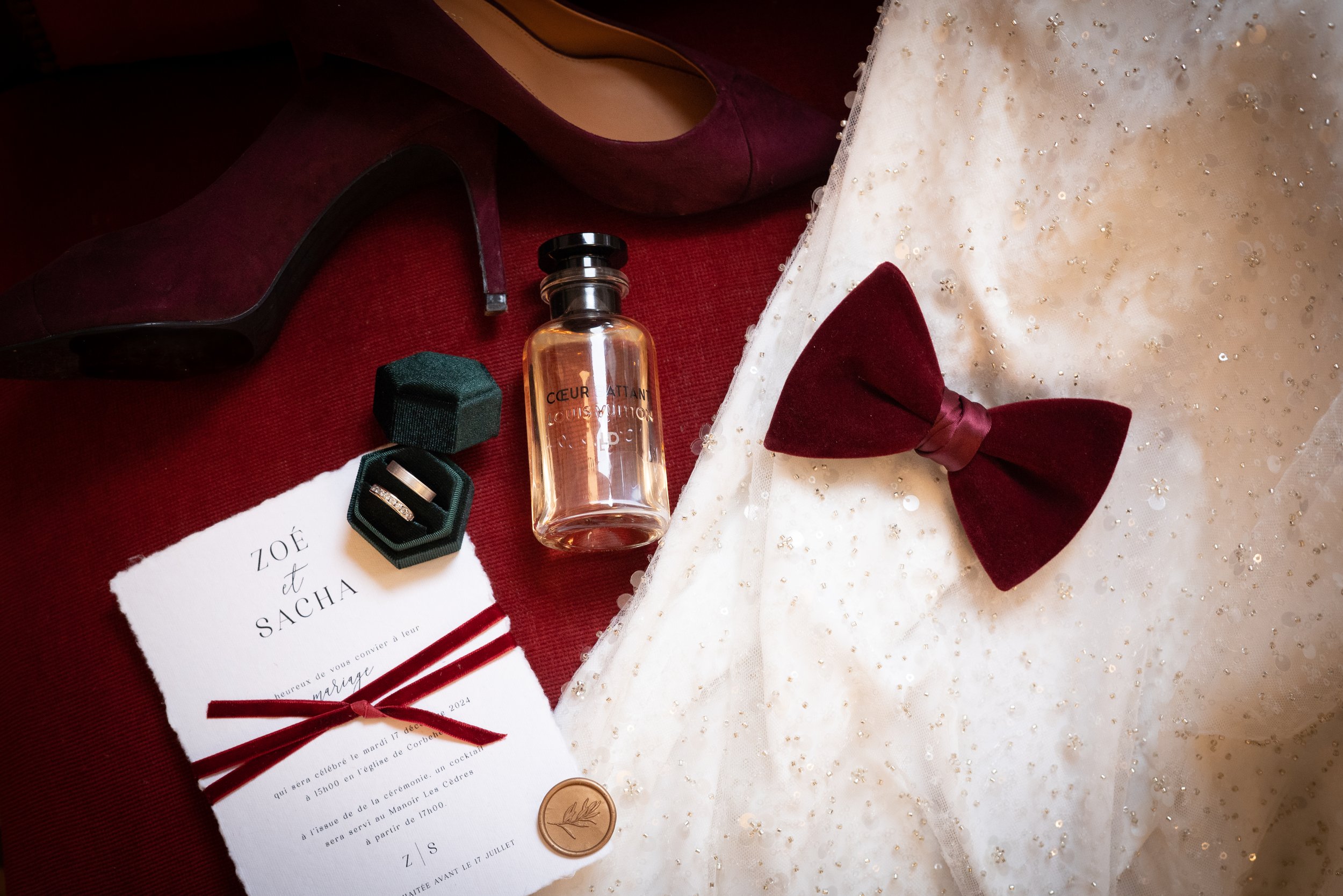 Accessoires de mariage comprenant une robe blanche à sequins, un nœud papillon bordeaux, une paire de chaussures bordeaux, un flacon de parfum, une alliance, une invitation de mariage, et une cire de sceau, tous disposés sur un fond en tissu rouge.