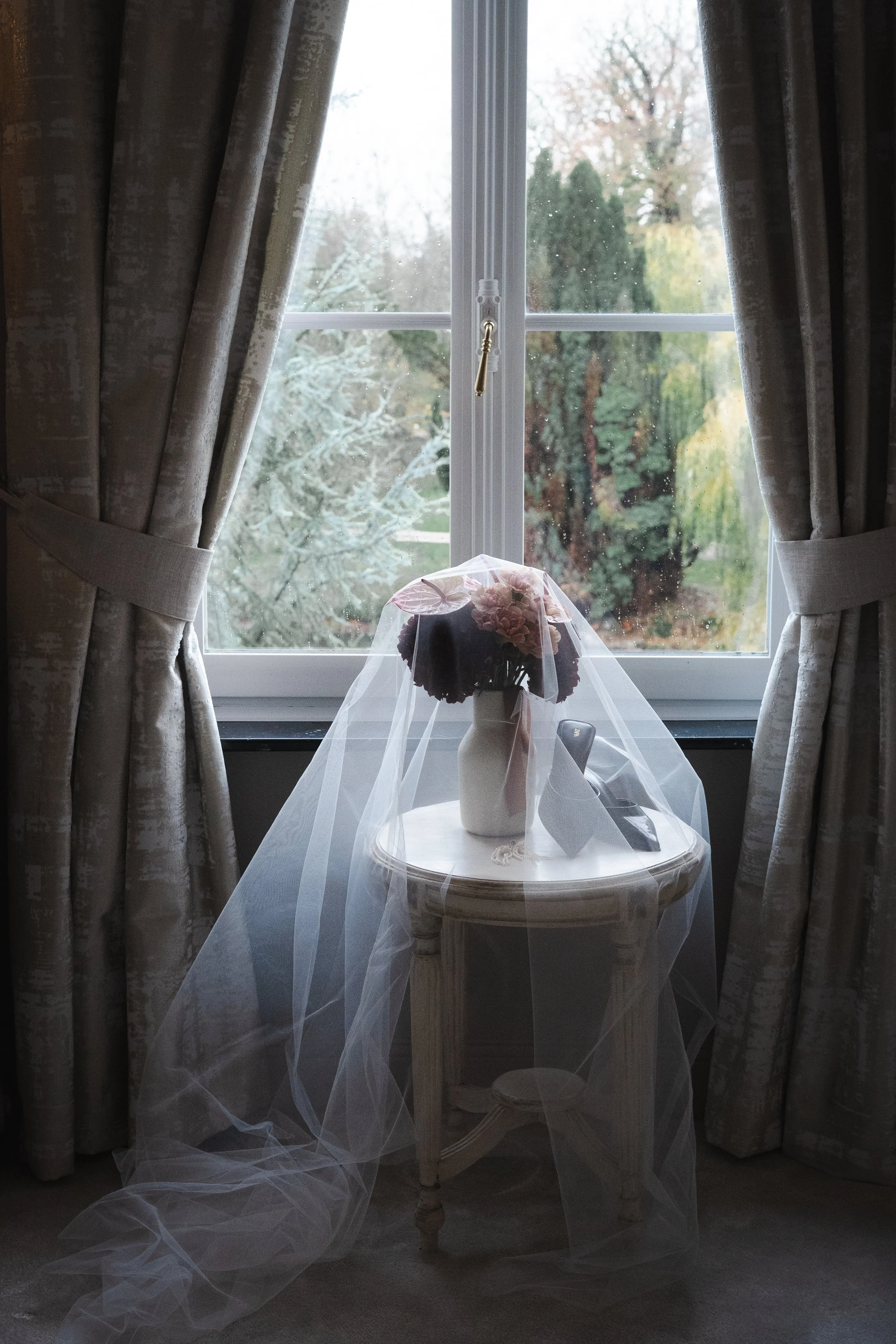 Une fenêtre avec des rideaux ouverts, une table ronde en bois blanc avec un vase contenant des fleurs, une télécommande, et un papier, un voile transparent est placé sur la table.