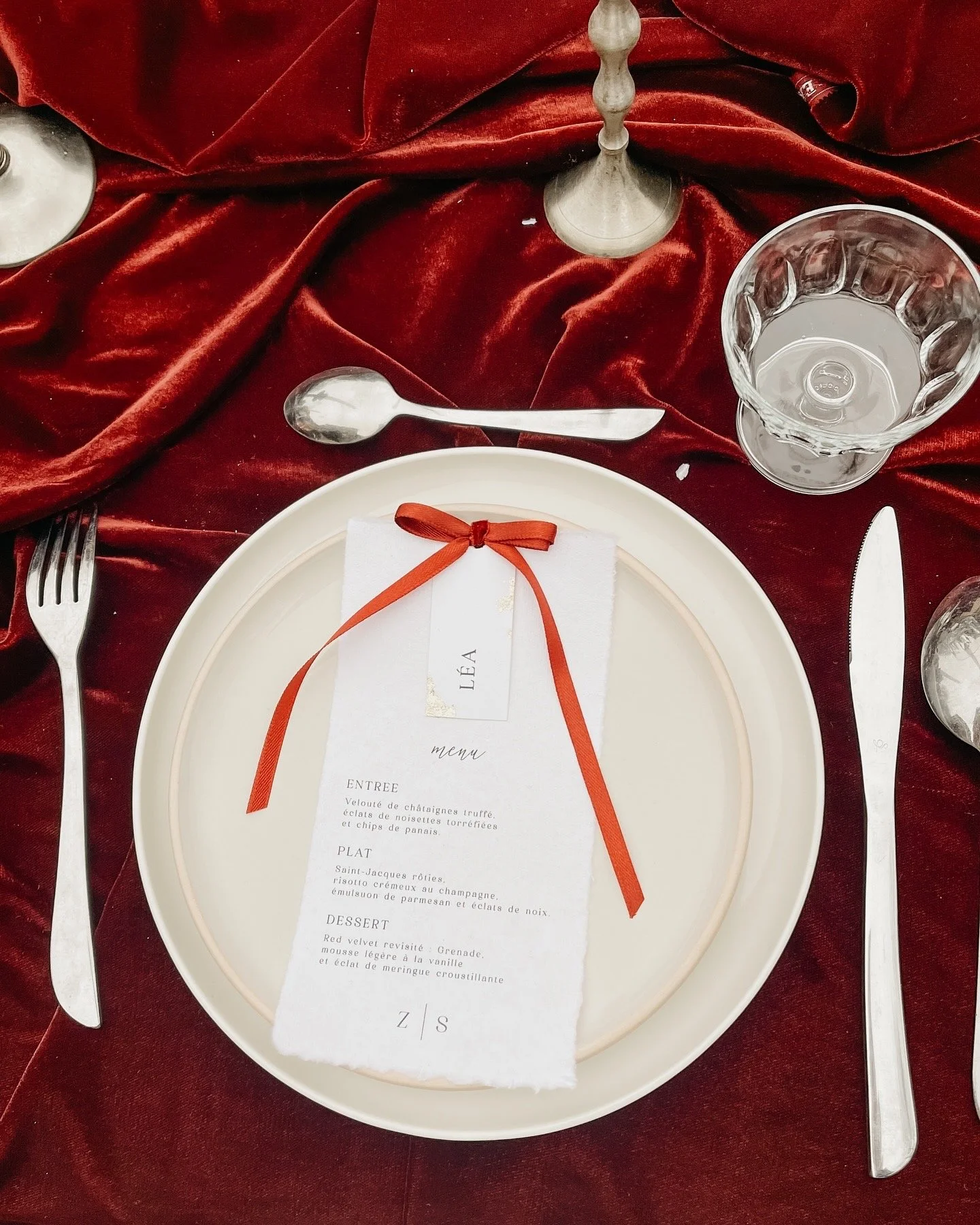 Une table élégante avec une assiette blanche, un menu en papier, un ruban rouge, une fourchette à gauche, un couteau à droite, un verre d'eau transparent, une cuillère en haut, et une nappe en velours rouge.