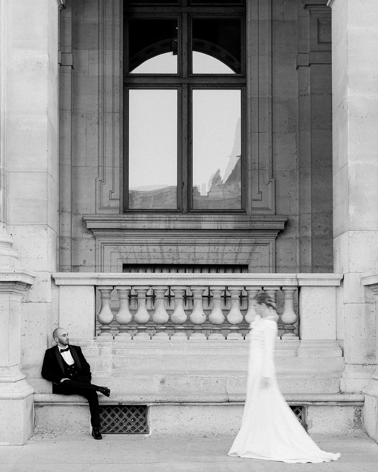 Photo en noir et blanc d'un homme en smoking assis sur un banc en pierre, à gauche, et d'une femme en robe de mariée passant devant lui, floue, à droite, devant une façade en pierre avec une grande fenêtre.