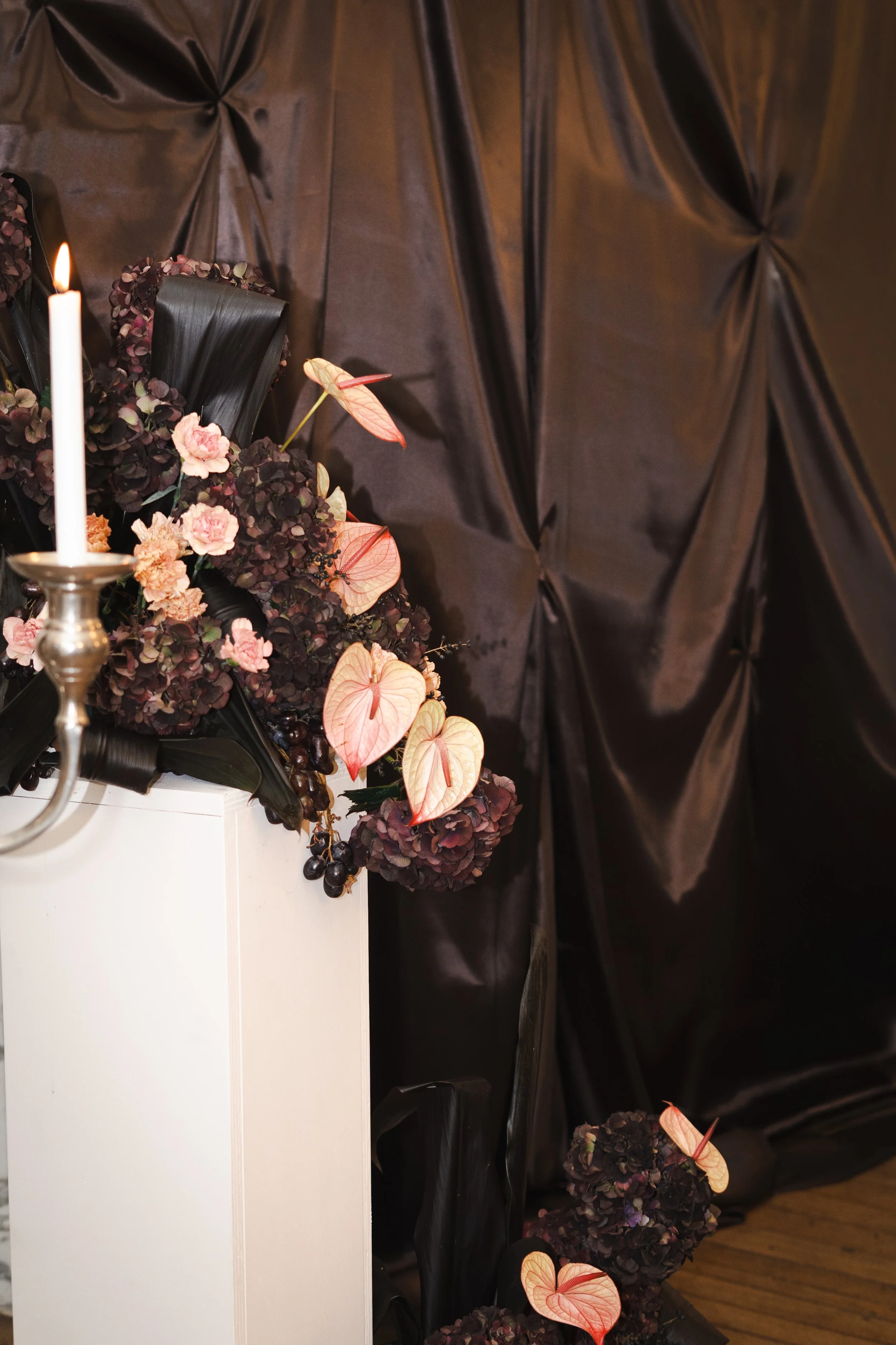 Décoration florale composée d'hortensias, d'anthuriums et d'éléments de verdure, avec un cadre noir en fond et une bougie allumée sur un support métallique.