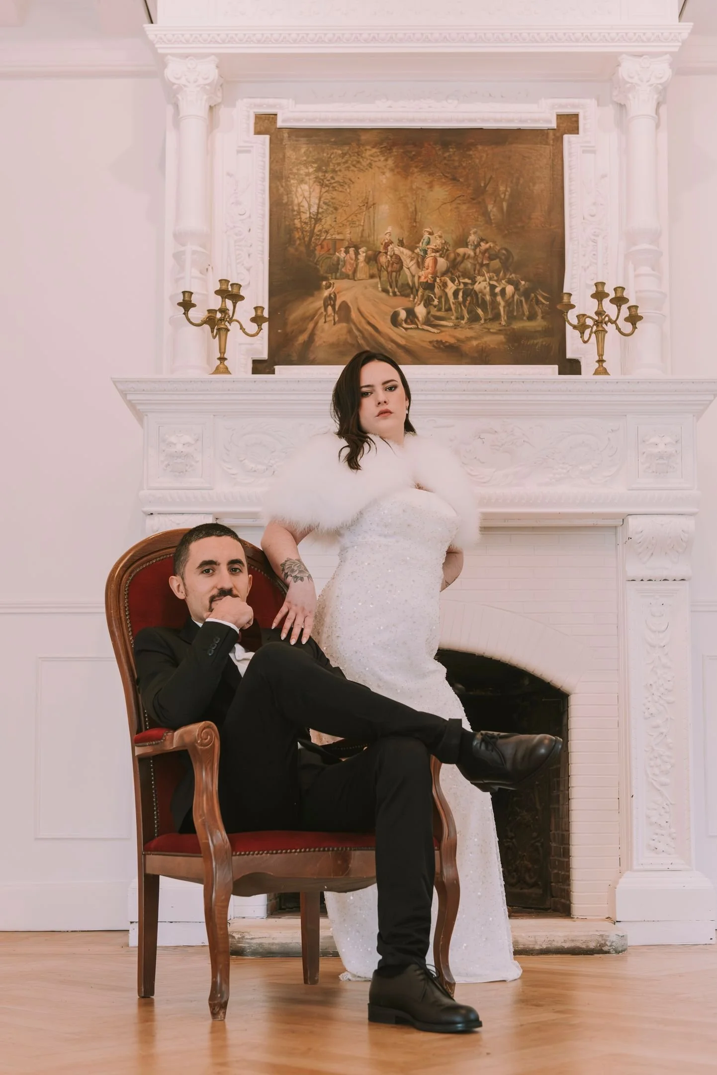 Une femme en robe de mariée blanche et un homme en smoking noir posent dans une pièce élégante avec une cheminée blanche et un tableau ancien au mur.
