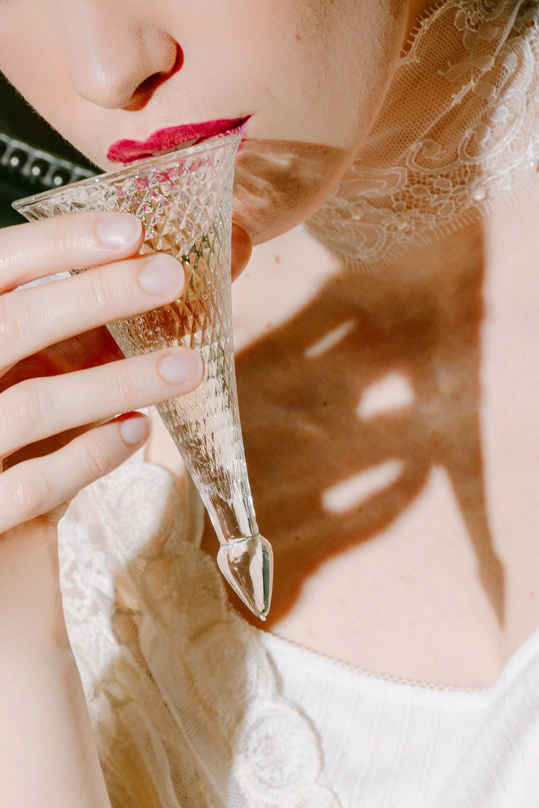 Une personne tient un verre à champagne en cristal, approchant le verre de ses lèvres peintes en rouge, avec un haut en dentelle blanche brodée.