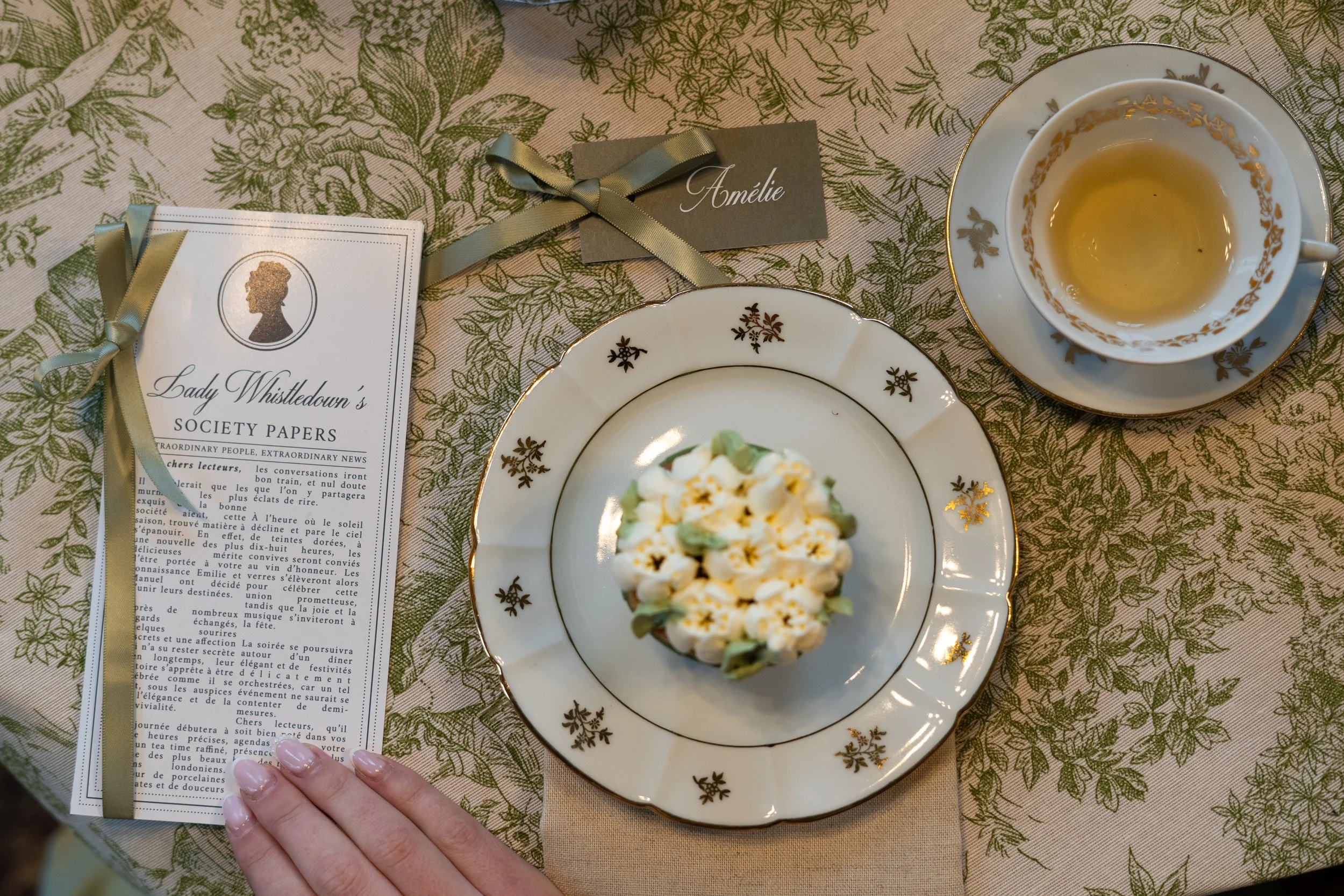 Une tasse de thé ou de tisane dans une tasse en porcelaine blanche avec des accents dorés, une assiette avec un cupcake décoré de petits fleurs blanches et de feuilles vertes, une feuille de serviette beige, une carte de nom "Amélie" attachée avec un