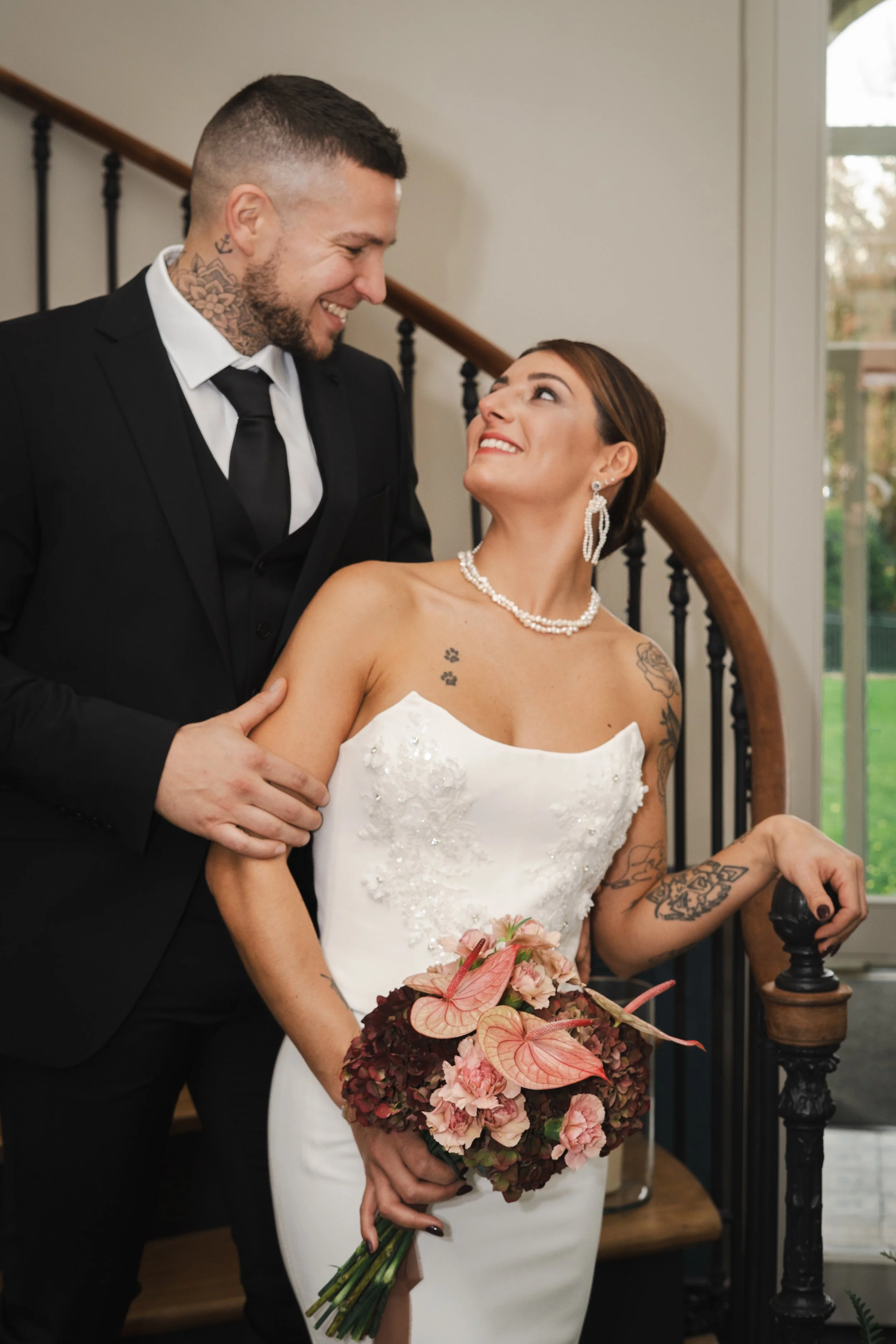 Un couple de mariés posant ensemble à l'intérieur, avec la mariée tenant un bouquet de fleurs roses et orange, la mariée en robe blanche et le marié en costume noir, se regardant avec amour.