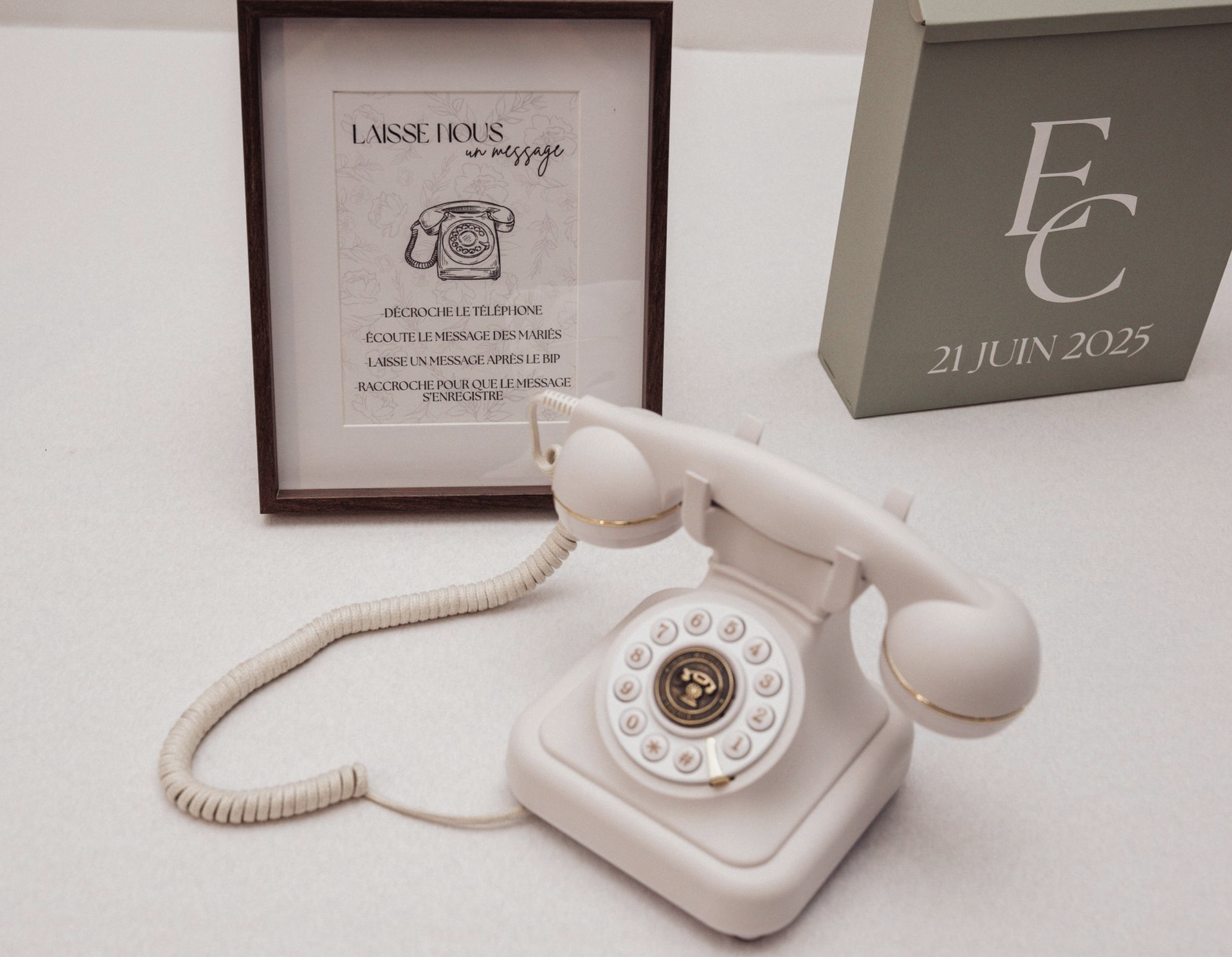 Téléphone vintage blanc avec cadran numéroté, placé sur une surface blanche, avec une affiche en arrière-plan contenant un dessin de téléphone et des instructions en français, et une boîte grisâtre avec des initiales et une date 21 juin 2025.