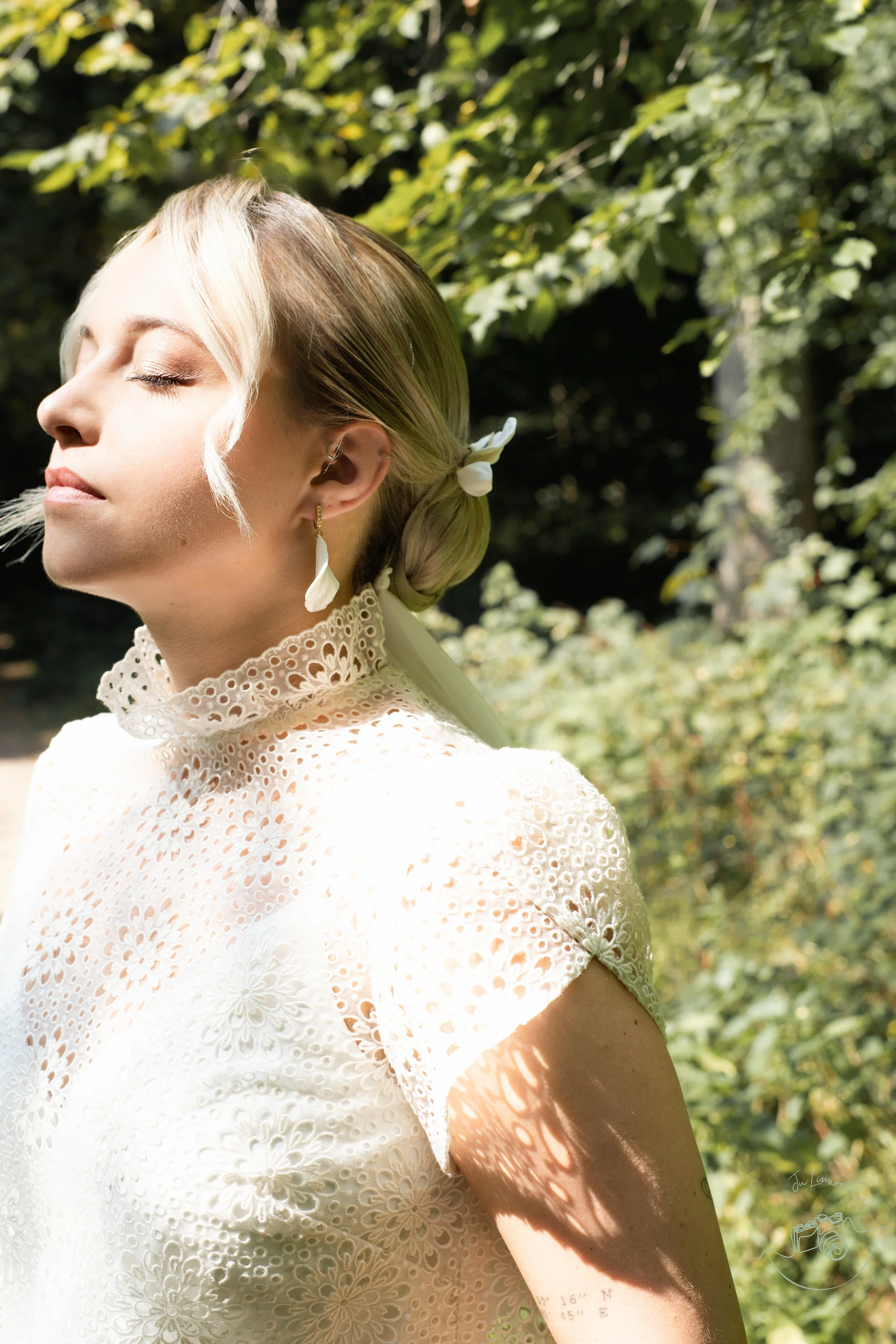 Femme avec visage détendu, portant une robe en dentelle blanche, bijoux, dans un environnement naturel ensoleillé.