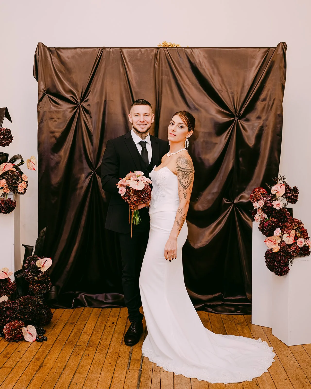 Un couple en tenue de mariage pose devant un fond en tissu marron avec des fleurs artificielles roses et marron sur les côtés.