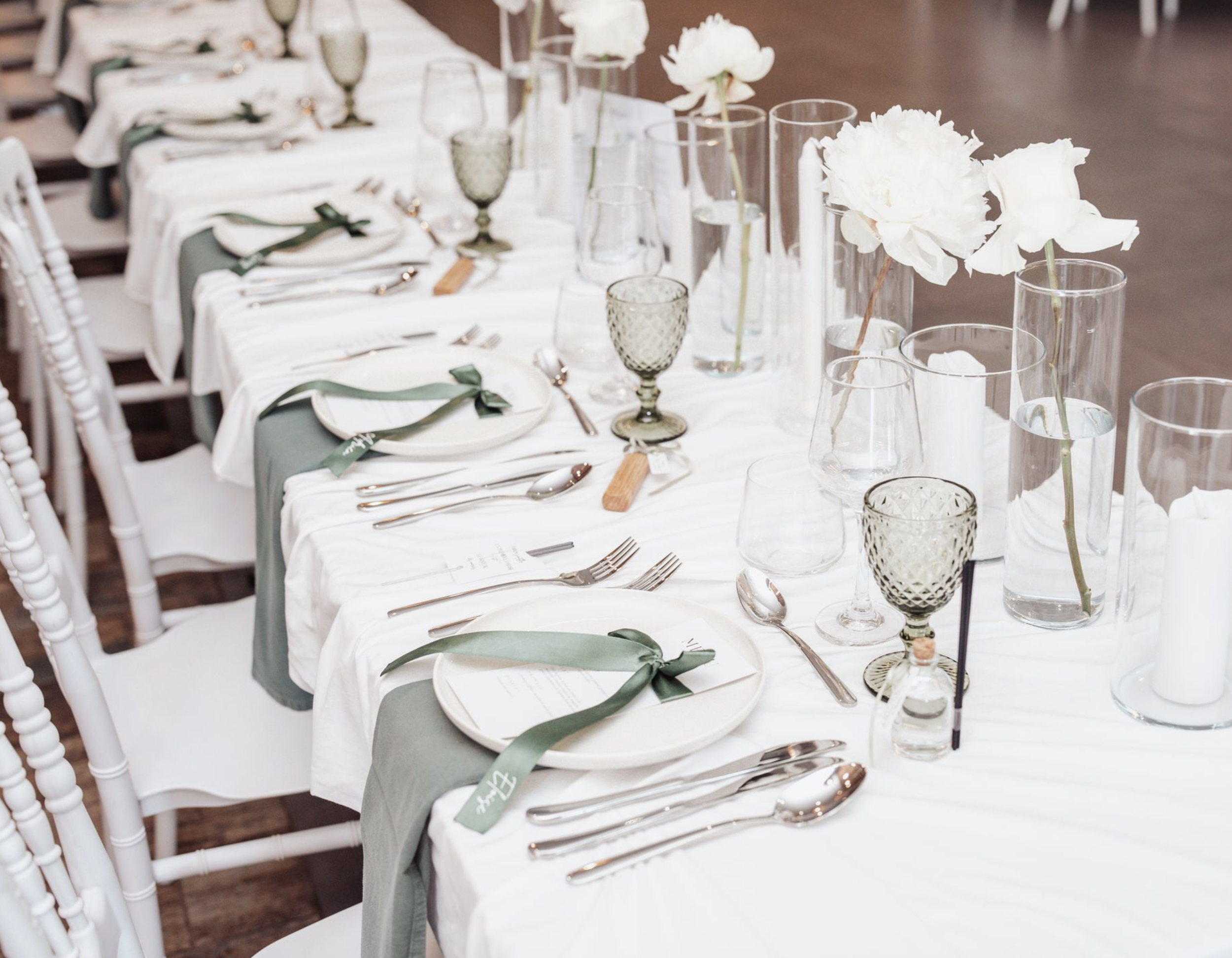 Table de mariage élégante avec vaisselle blanche, couverts en argent, verres à vin, fleurs blanches dans des vases et serviettes vertes.
