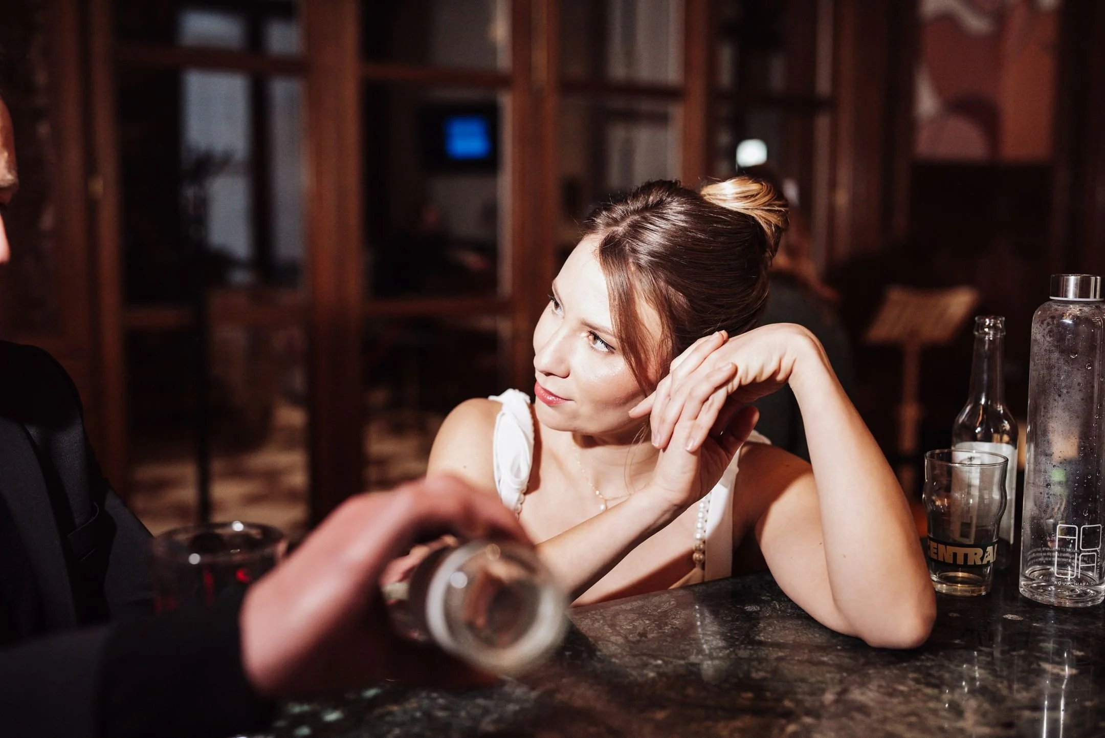 Une femme assise à une table en bois dans un bar ou un restaurant, elle regarde vers la gauche, avec les mains soutenant sa tête. Il y a des bouteilles et un verre sur la table.