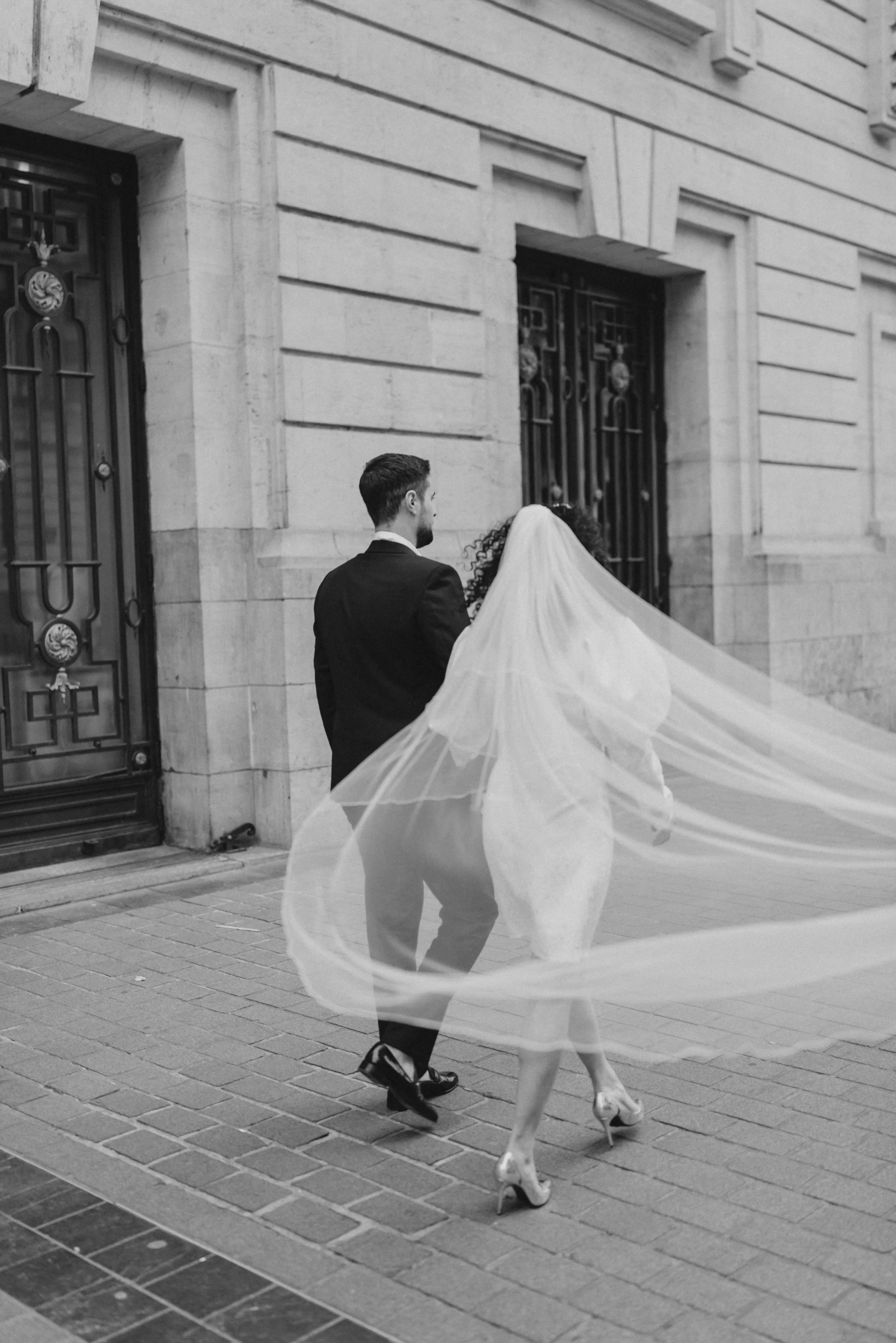 Un couple de mariés, vu de dos, marchant dans une rue pavée devant un bâtiment en pierre, la femme portant une robe de mariée et un voile longue, en talons hauts, et l'homme en costume noir.