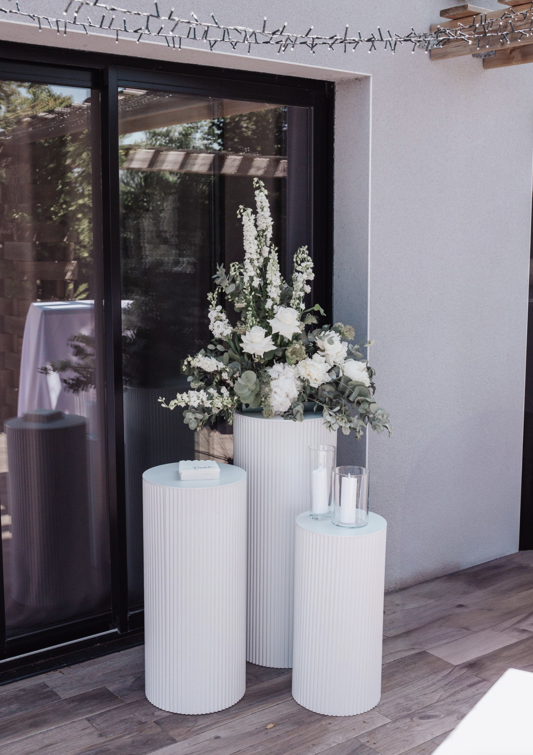Un grand vase blanc avec un arrangement floral de fleurs blanches, deux petits vases avec des bougies, sur trois piédestals blancs rayés, à l'extérieur d'une maison avec une porte en verre.