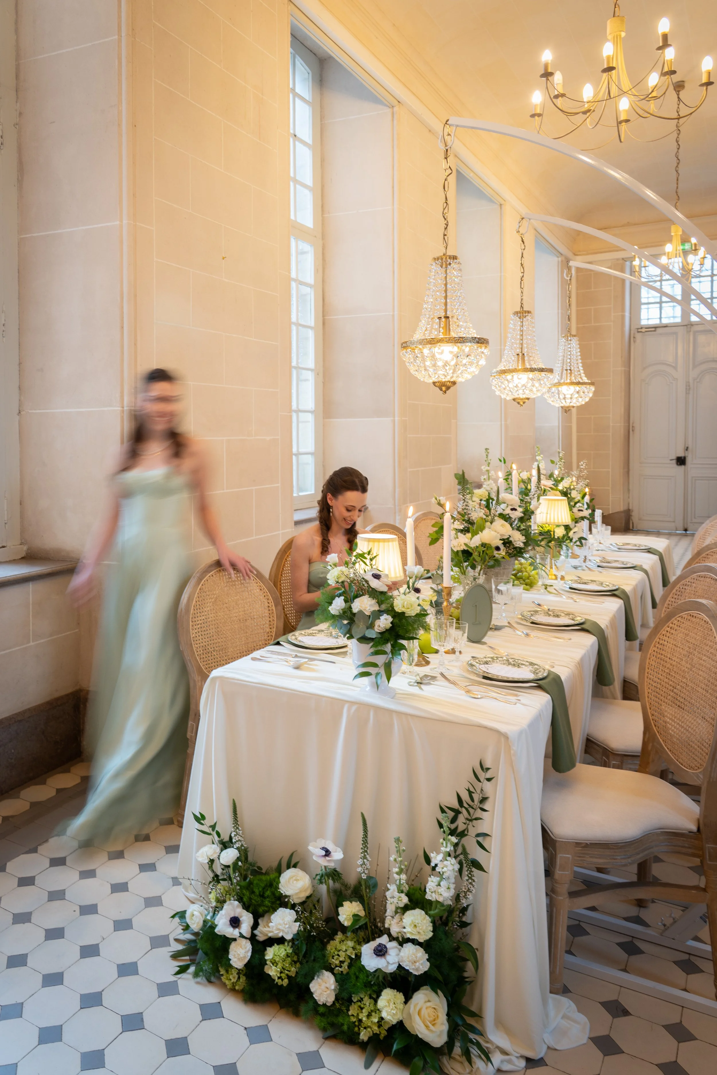 Une salle de banquet élégante avec une longue table décorée de fleurs blanches, chandelles et vaisselle, dans un intérieur lumineux avec grands fenêtres et lustres en cristal, où une femme est assise et une autre se tient à côté.