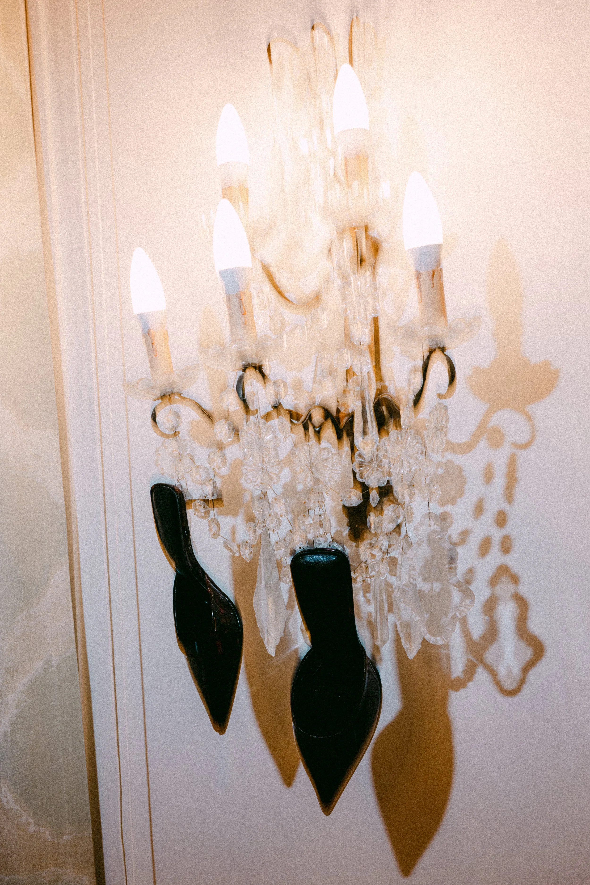 Un lustre en cristal avec cinq appliques, deux chaussures noires suspendues en dessous, et leur ombre projetée sur le mur.