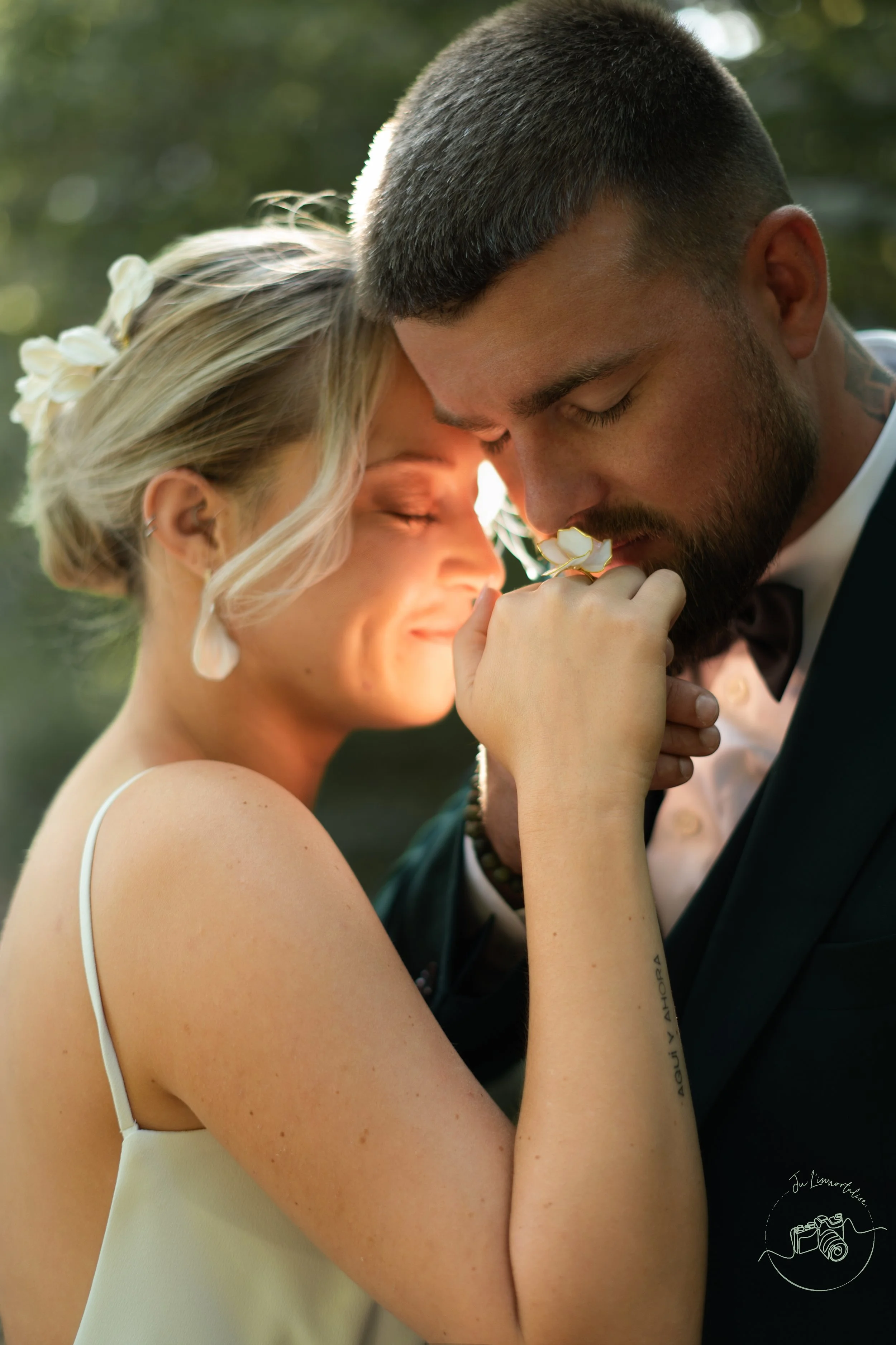 Un couple se tient près l'un de l'autre, la femme sourit avec les yeux fermés, tenant une fleur à la main, en tenue de mariage, dans un environnement naturel.