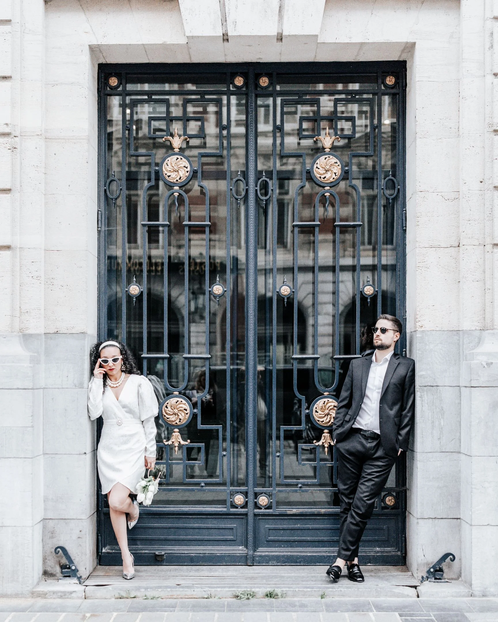 Deux personnes élégantes posant devant une grande porte en fer forgé décorée en or sur un bâtiment en pierre.
