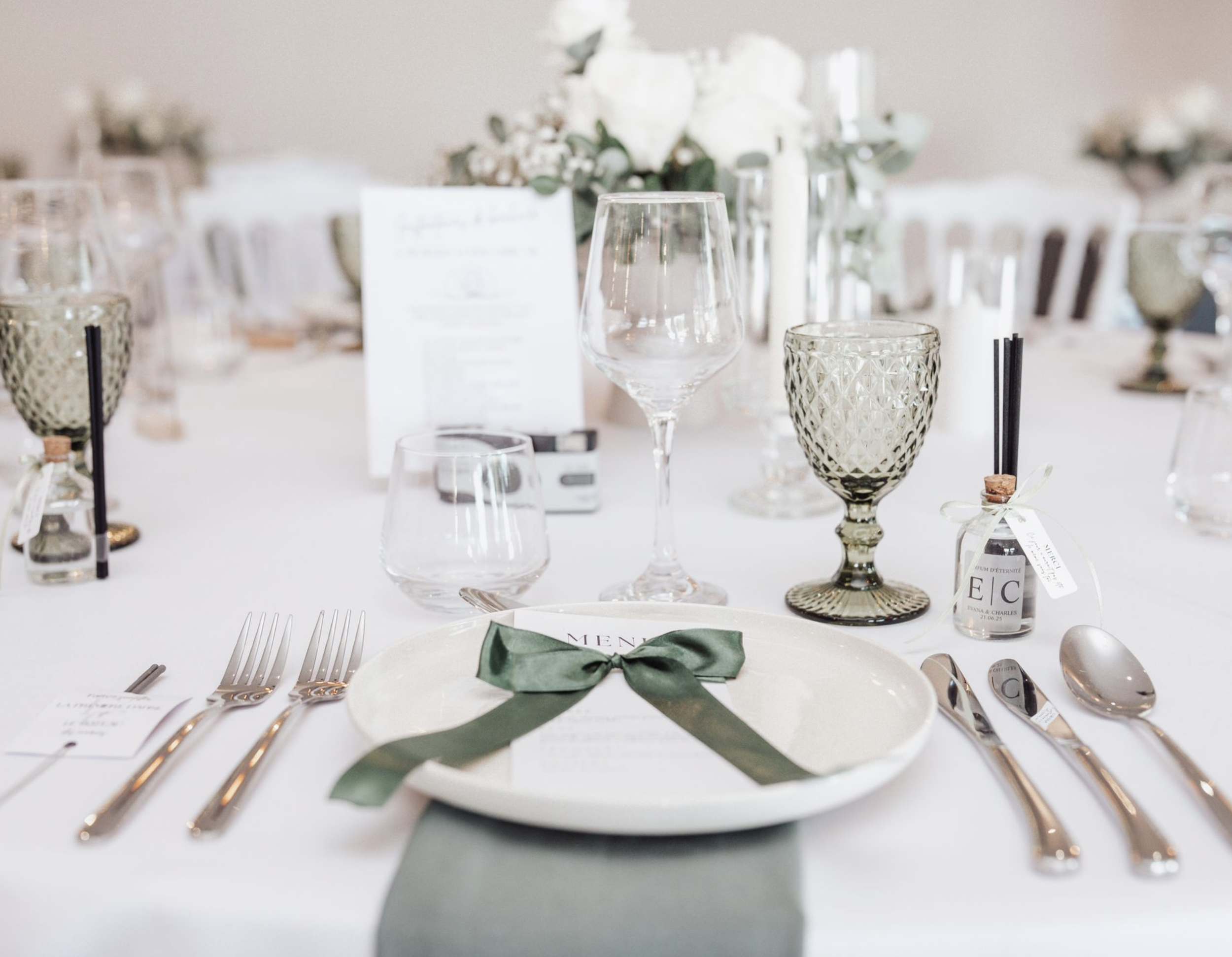 Table de mariage élégante avec vaisselle, couverts, verres, décoration florale blanche, livre de menu avec ruban vert.