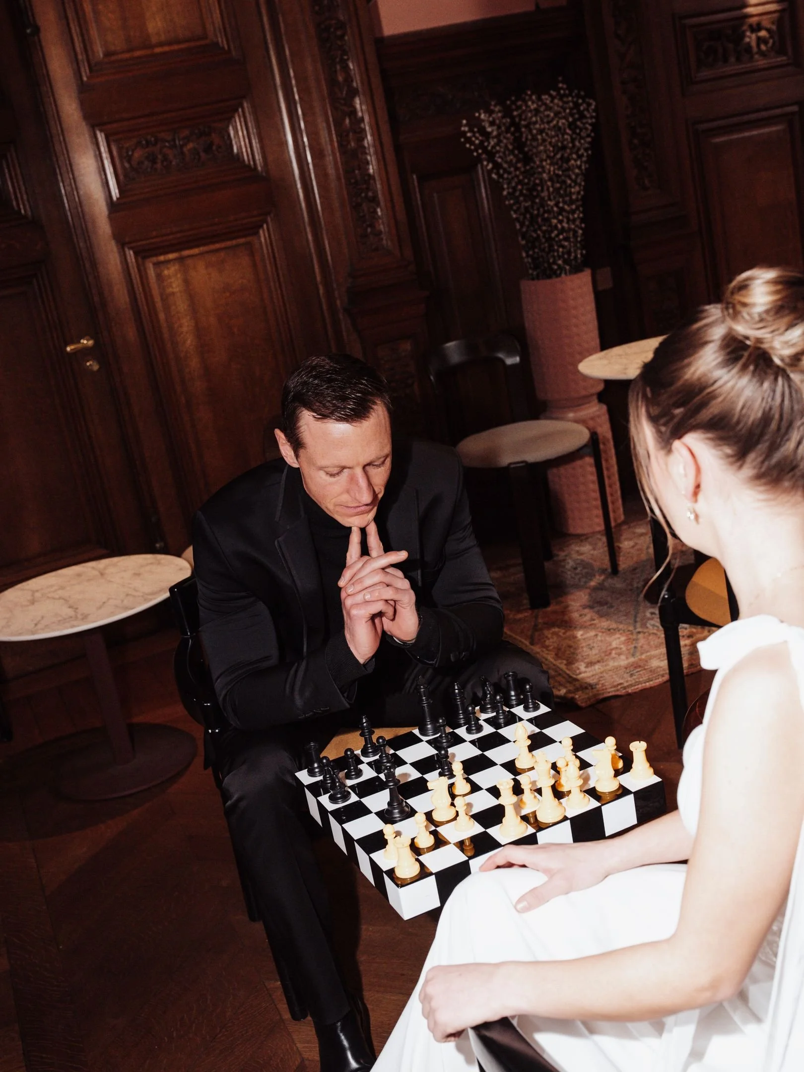 Deux personnes jouent aux échecs dans une pièce avec un décor en bois, un homme en costume noir et une femme en robe blanche, assis face à face.