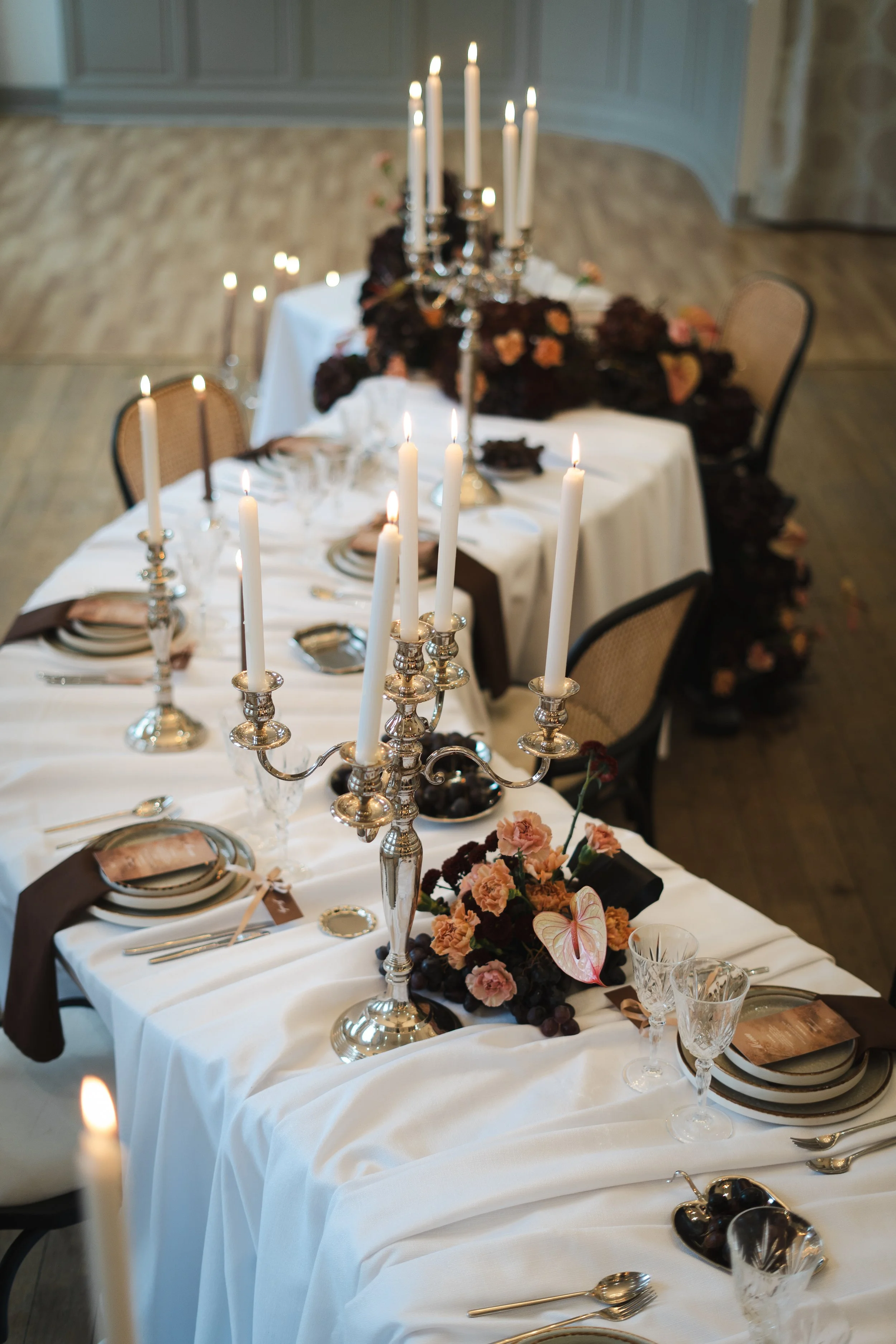 Table de fête élégante avec une nappe blanche, des chandeliers en argent et des bougies allumées, décorée de fleurs roses, noires et orange, prête pour un dîner sophistiqué.